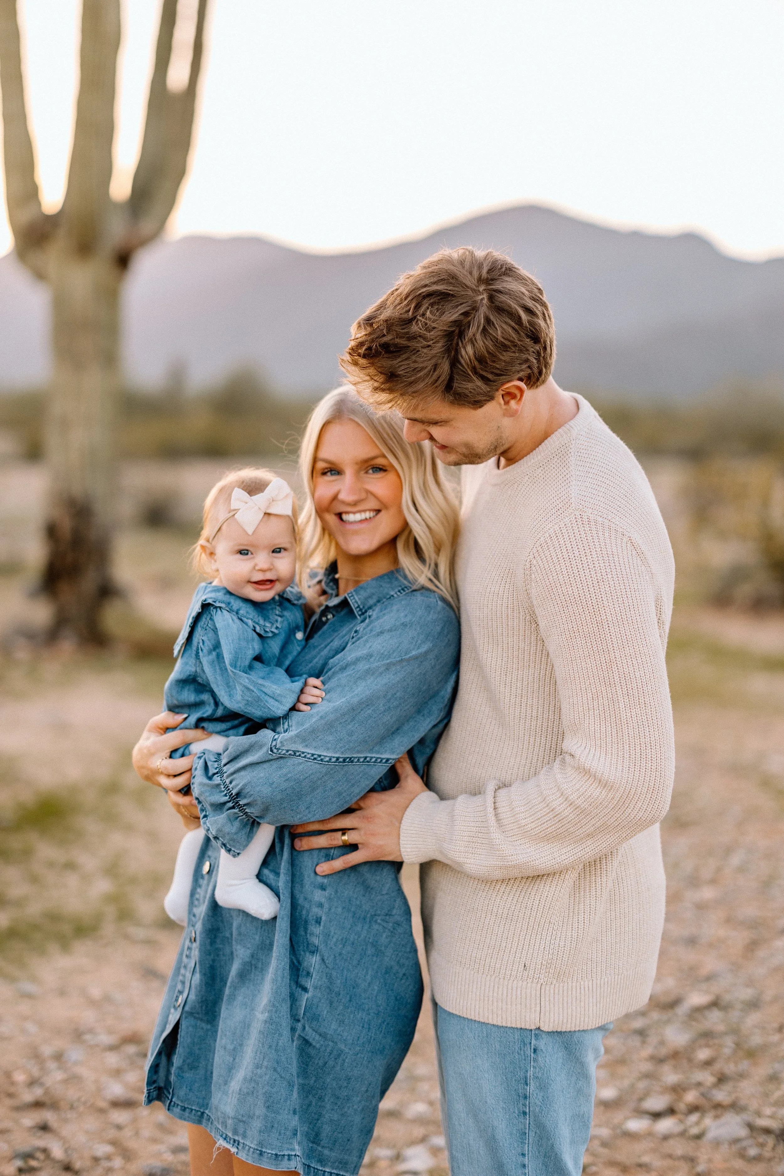 A family of three standing outdoors in a desert landscape with mountains in the background. The woman is holding a smiling toddler girl in her arms and the man is standing close beside them, looking down at the girl.