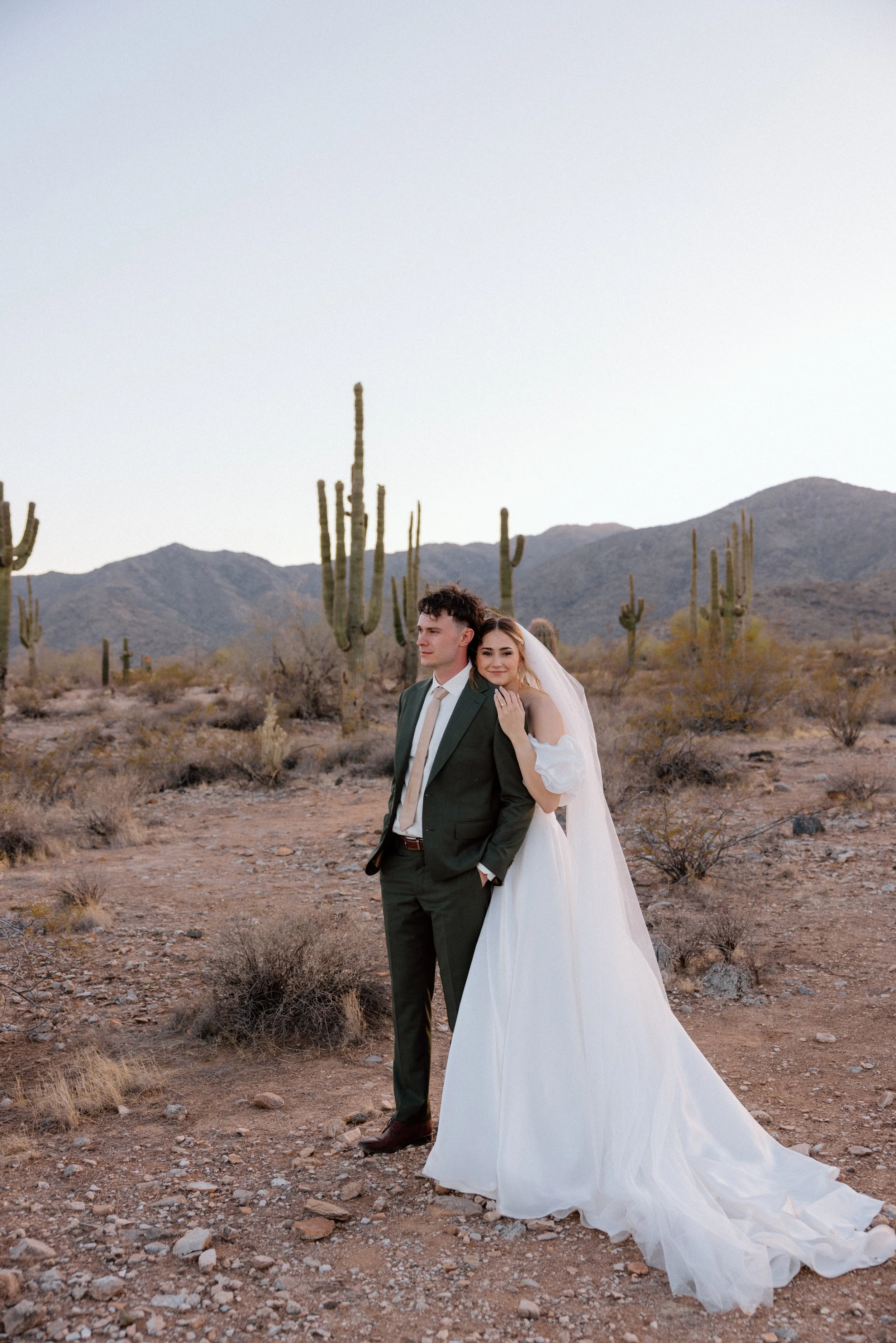 A bride and groom stand in a desert landscape with cacti and mountains in the background, during a wedding photoshoot.