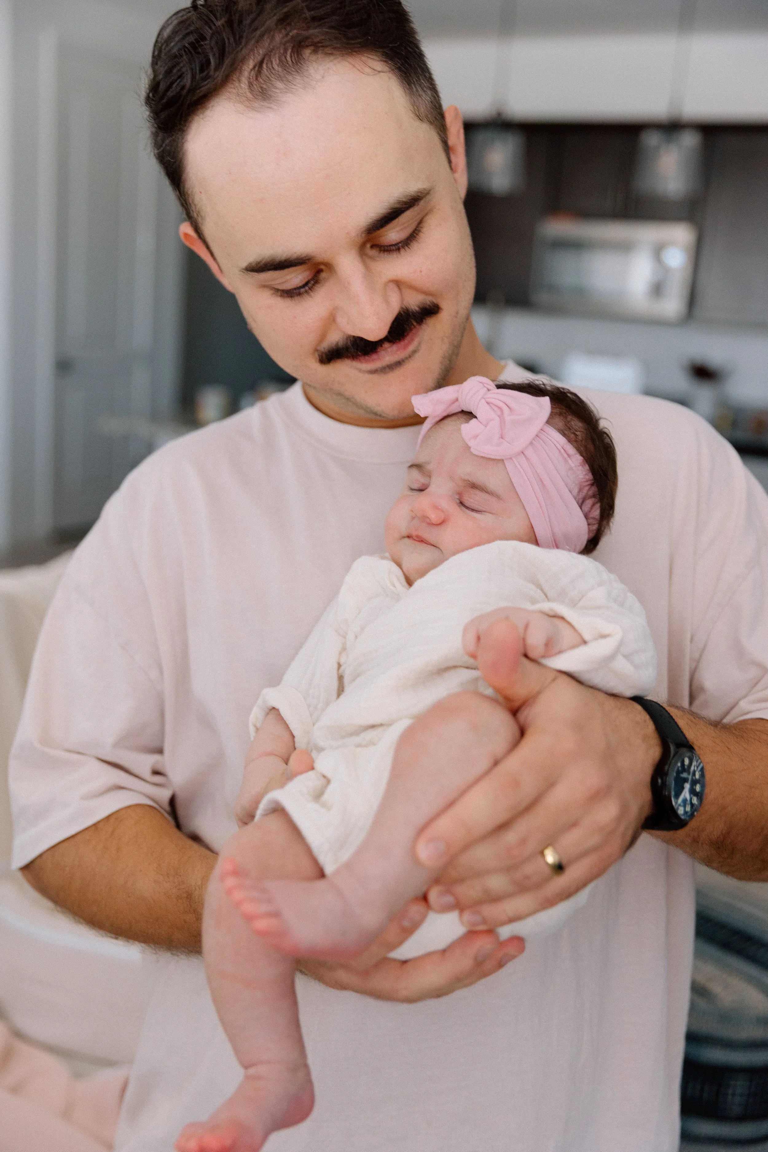 A man with a mustache holding a sleeping baby girl with a pink headband and white outfit in a home kitchen.