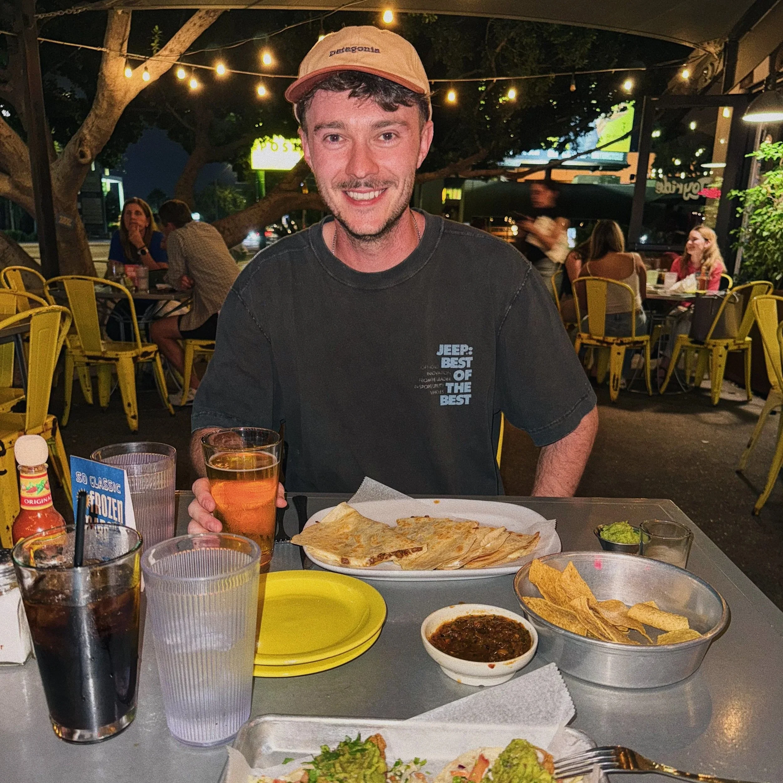 A young man smiling at a restaurant table at night, with food including quesadillas, chips, salsa, and drinks, in an outdoor setting with string lights and other diners in the background.