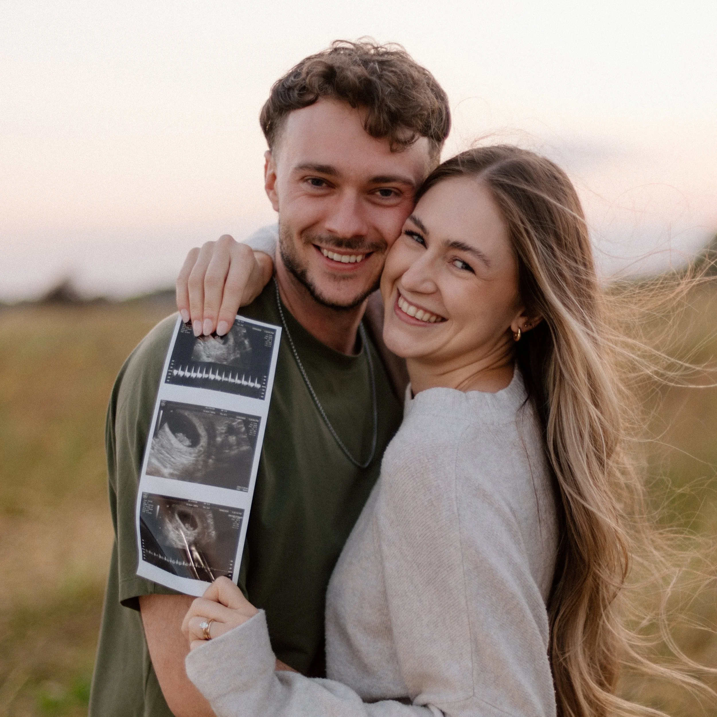 A couple smiling and hugging outdoors at sunset, with the man holding an ultrasound photo.
