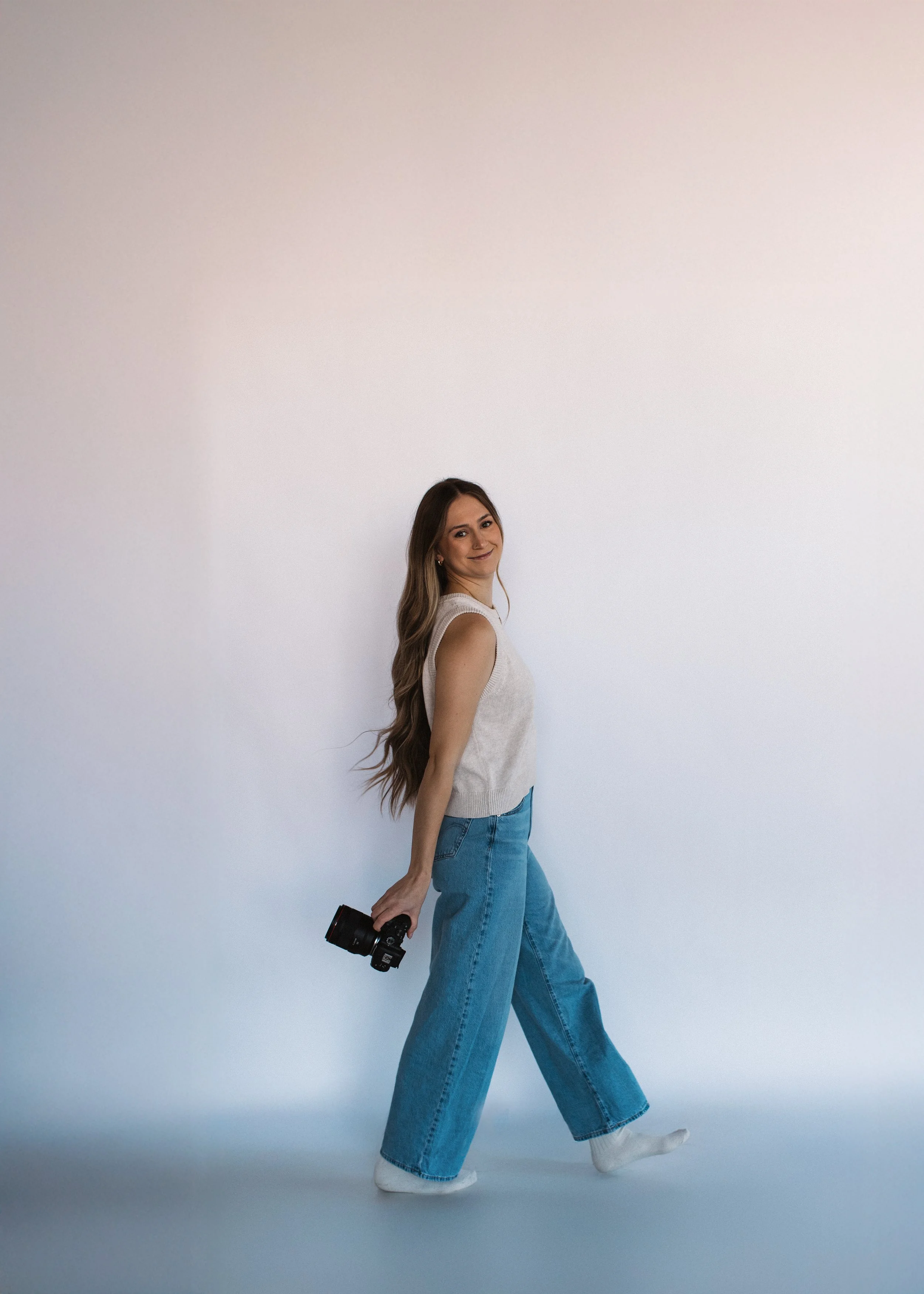 A young woman with long wavy hair dressed in a sleeveless white top and loose blue jeans, holding a camera in her right hand, walking against a plain, light-colored background, smiling at the camera.