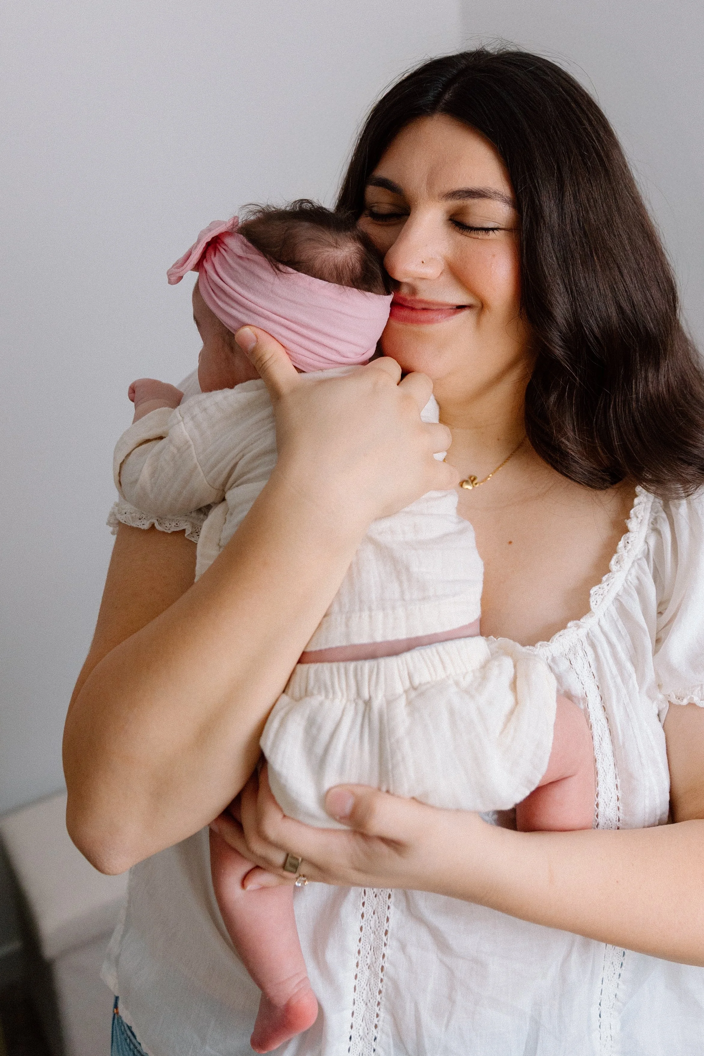 A woman holding a newborn baby, gently hugging and smiling, with the baby wearing a pink headband.