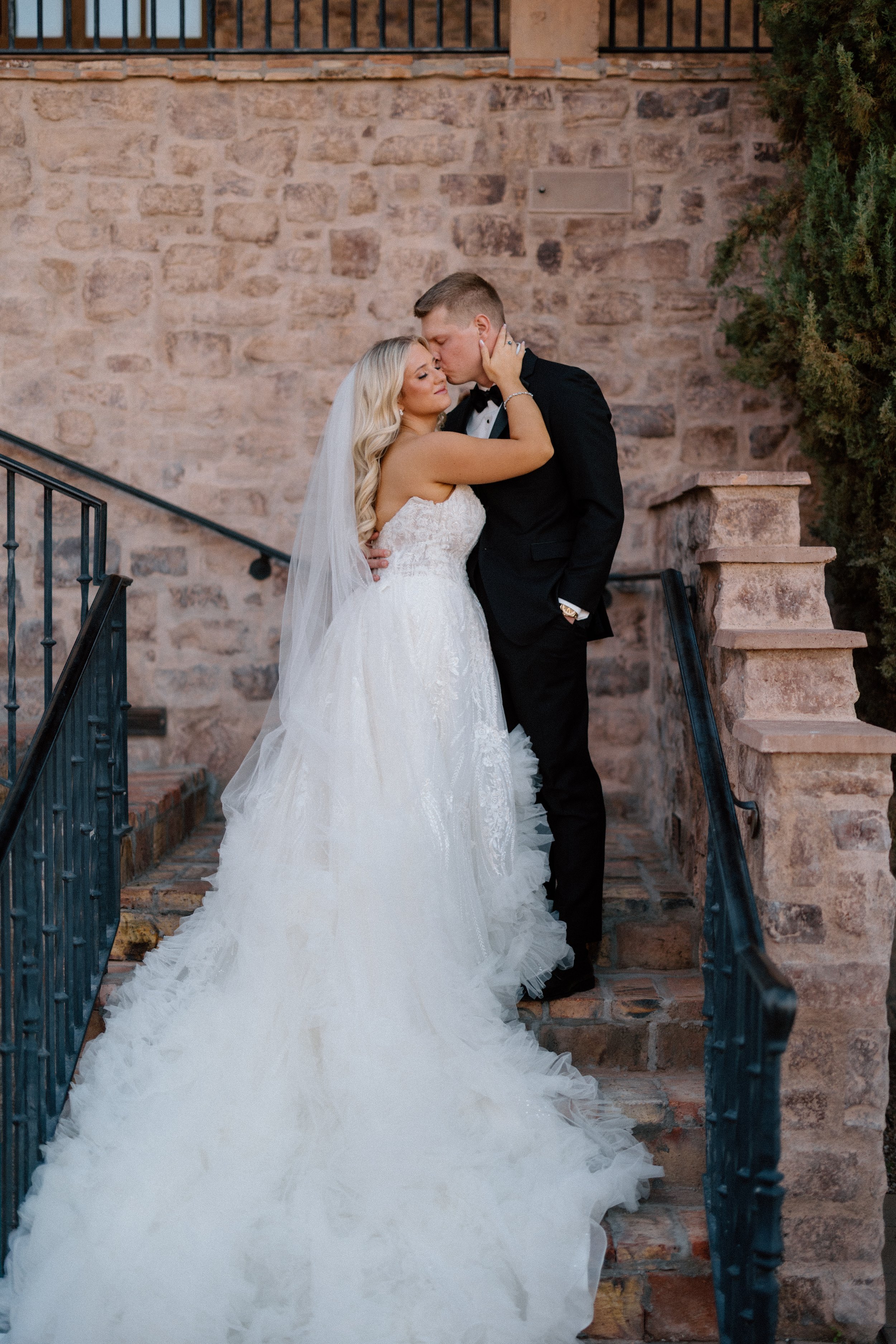 Bride and groom share an intimate moment on the stairs outdoors, with the bride in a white wedding gown and veil, and the groom in a black tuxedo, against a brick wall background.