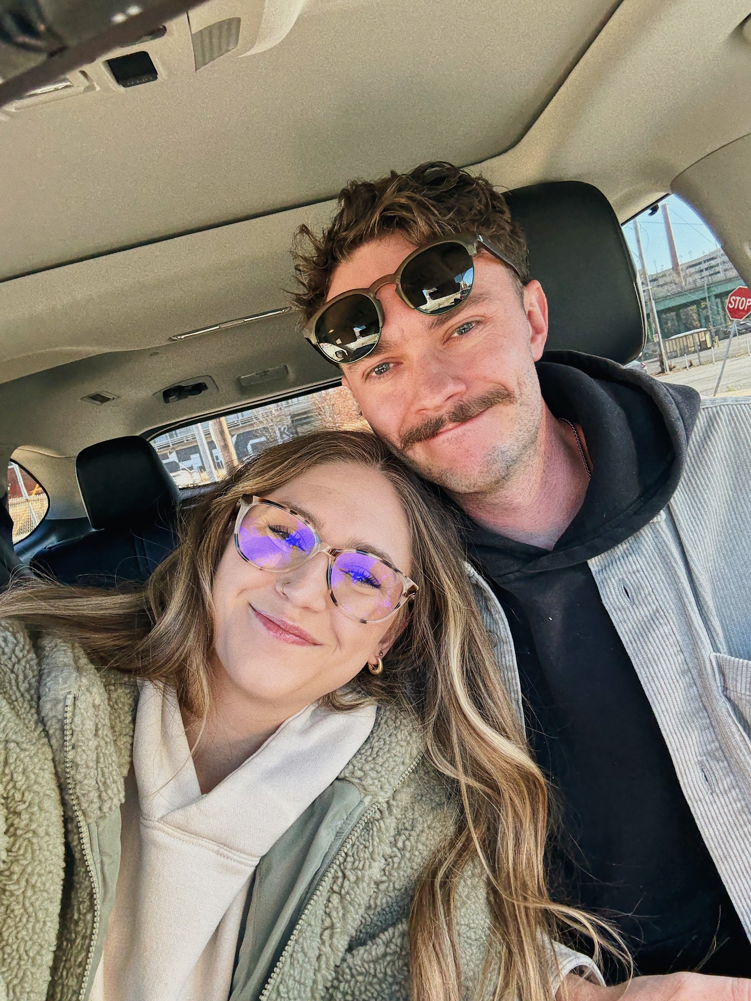 A young woman with glasses smiling and a man with sunglasses sitting close together inside a car.