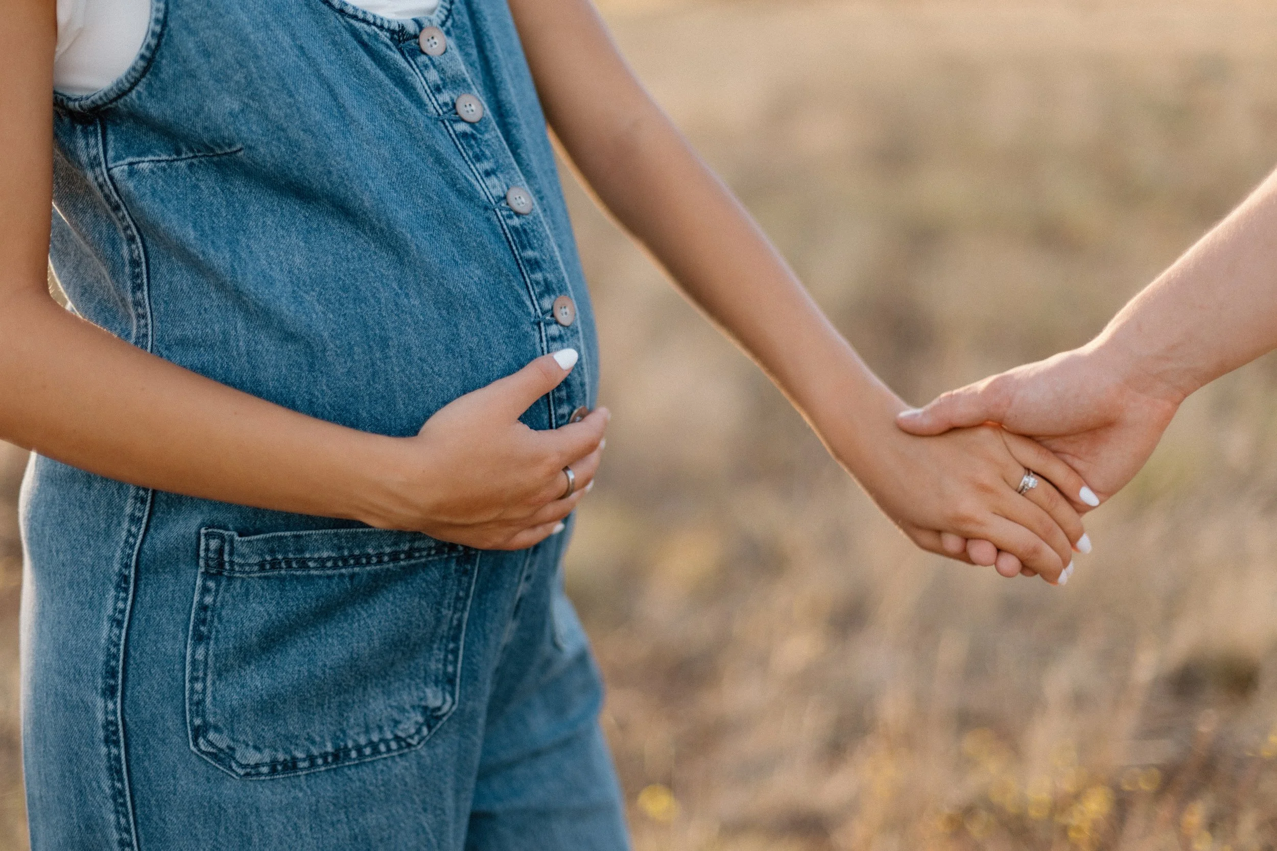 A pregnant woman in denim overalls holding her belly with one hand and holding hands with a partner with the other, outdoors in a natural setting.