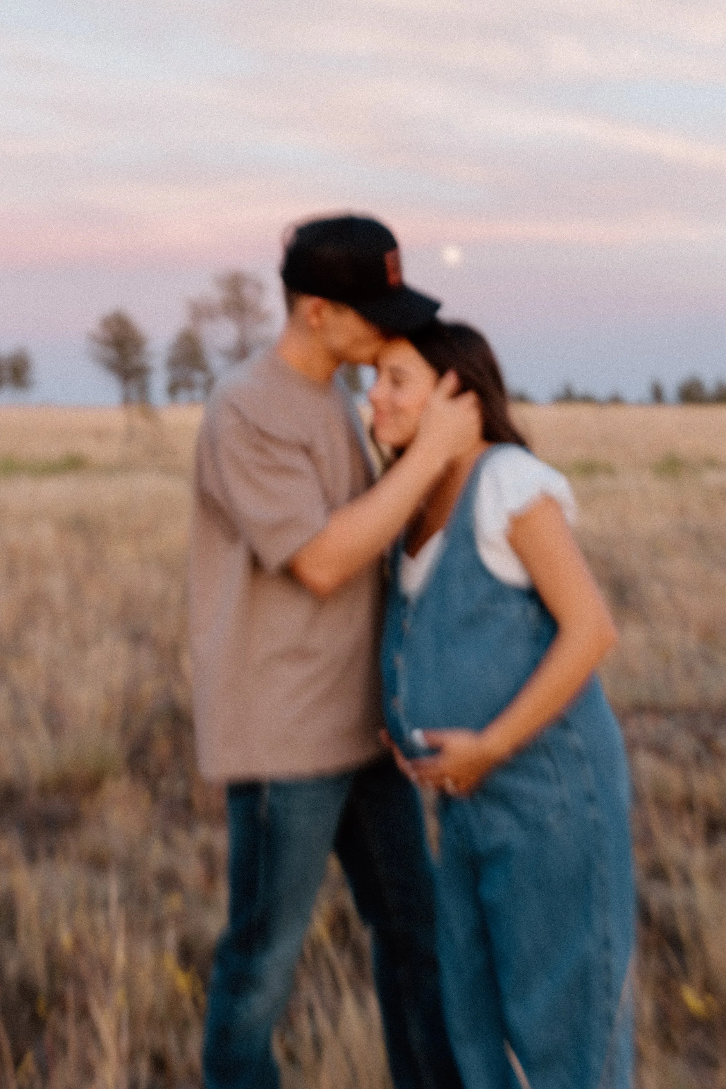 A couple standing close in a field during sunset, with the man gently touching the woman's face and the woman resting her hand on her pregnant belly.