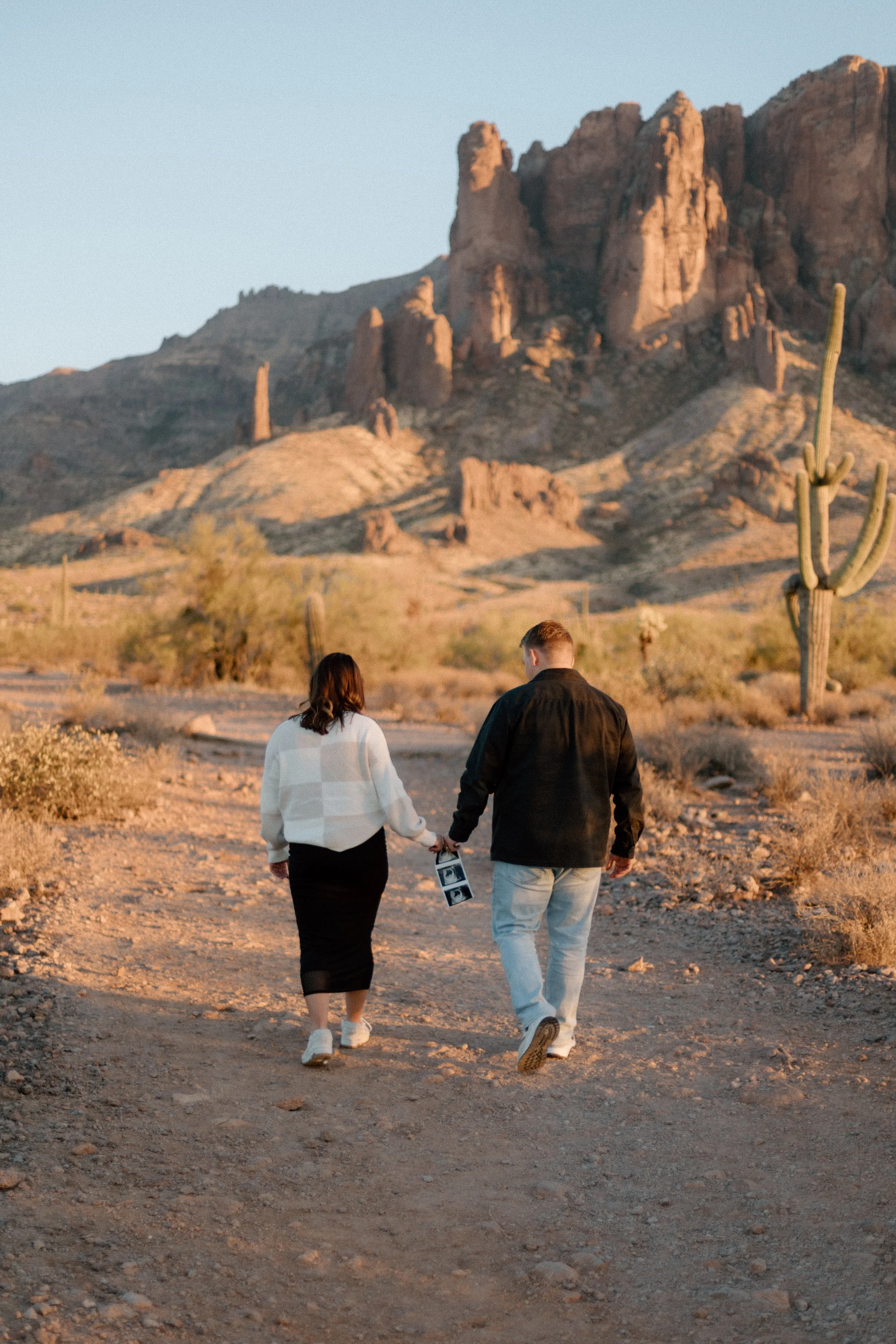 A couple walking on a dirt trail in a desert landscape with cacti and towering rocks in the background at sunset.