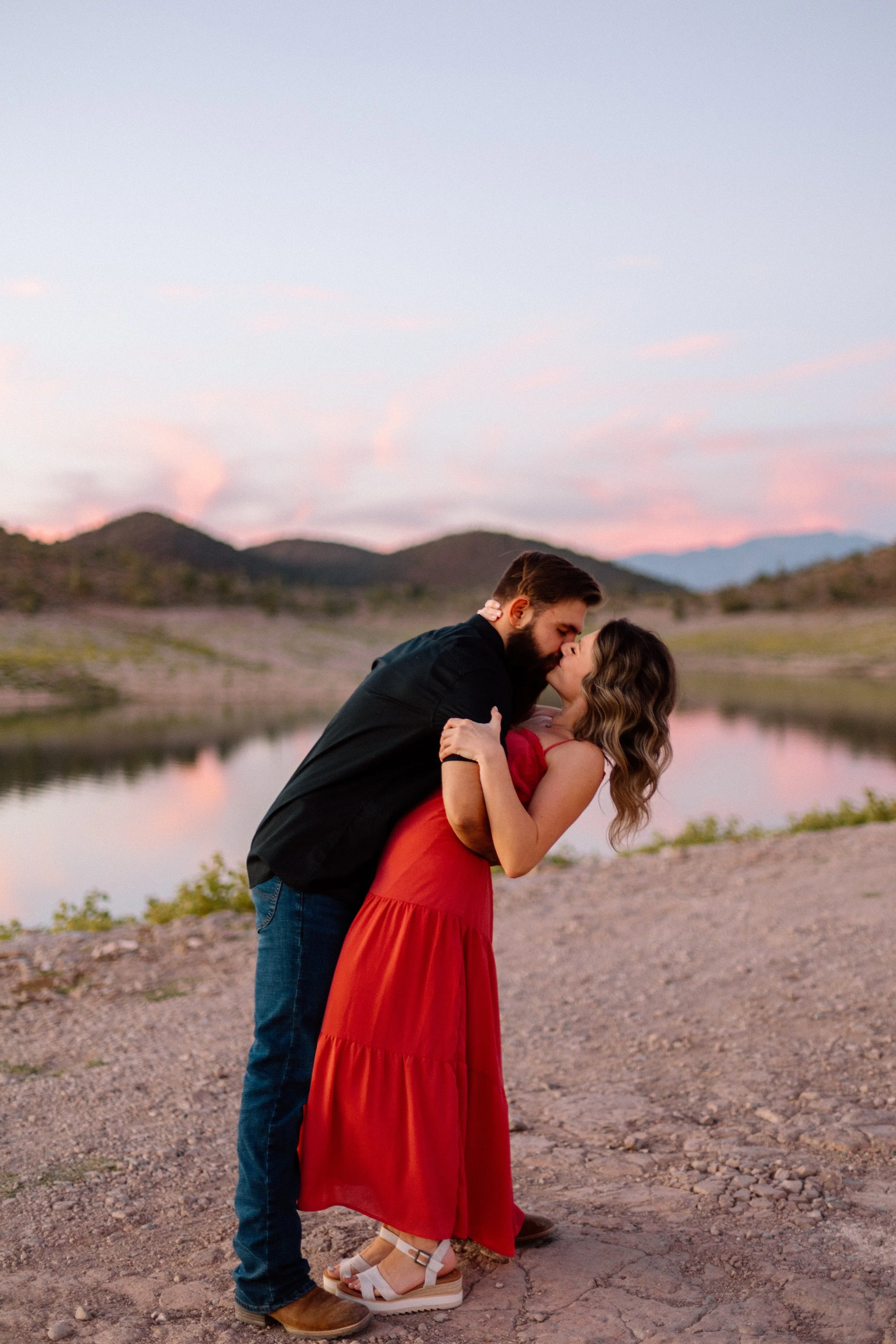 A couple kissing outdoors near a body of water at sunset, with mountains in the background.