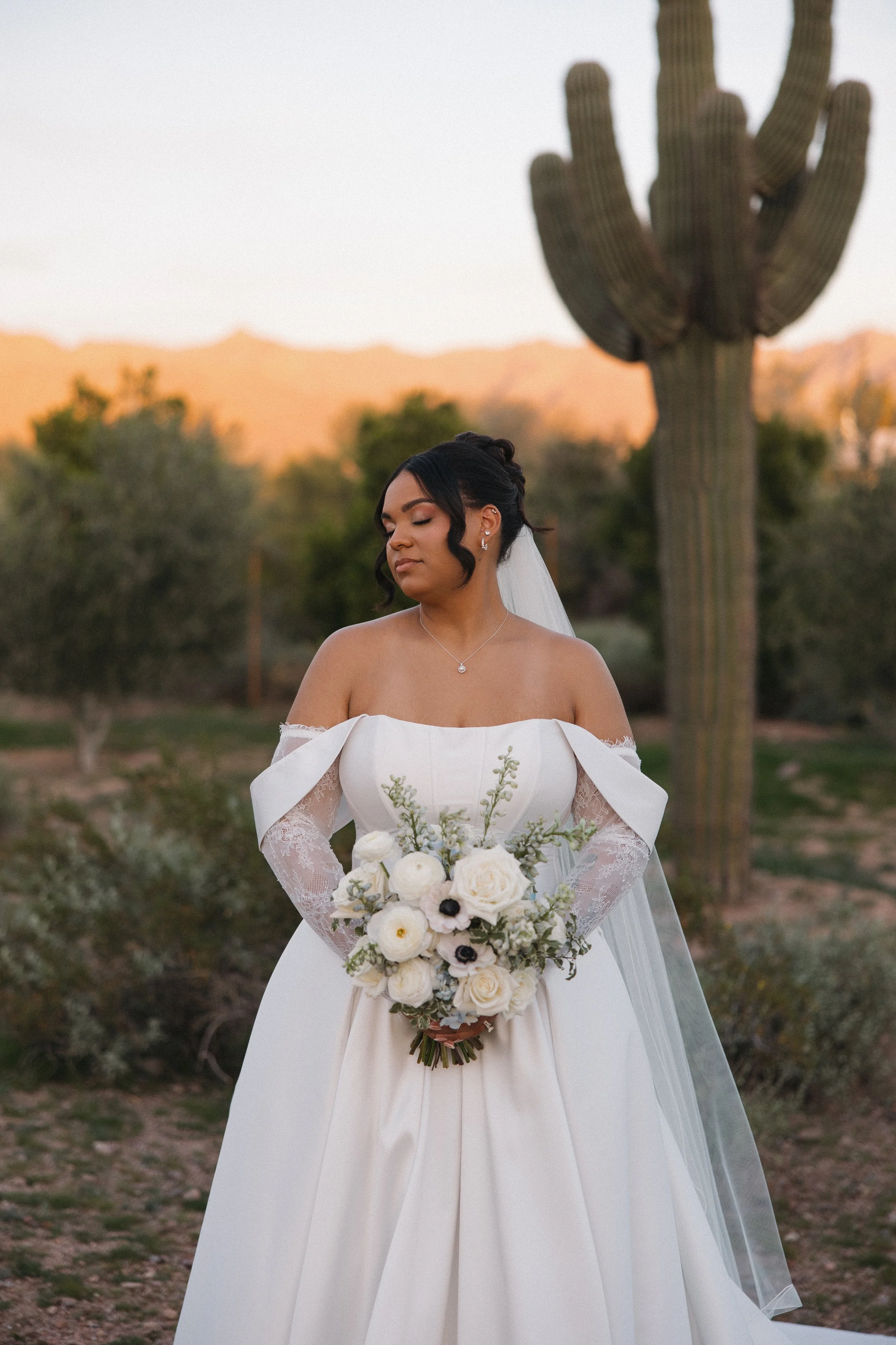 Bride in a white wedding gown holding a bouquet of white and dark purple flowers, standing outdoors with desert plants and mountains in the background.