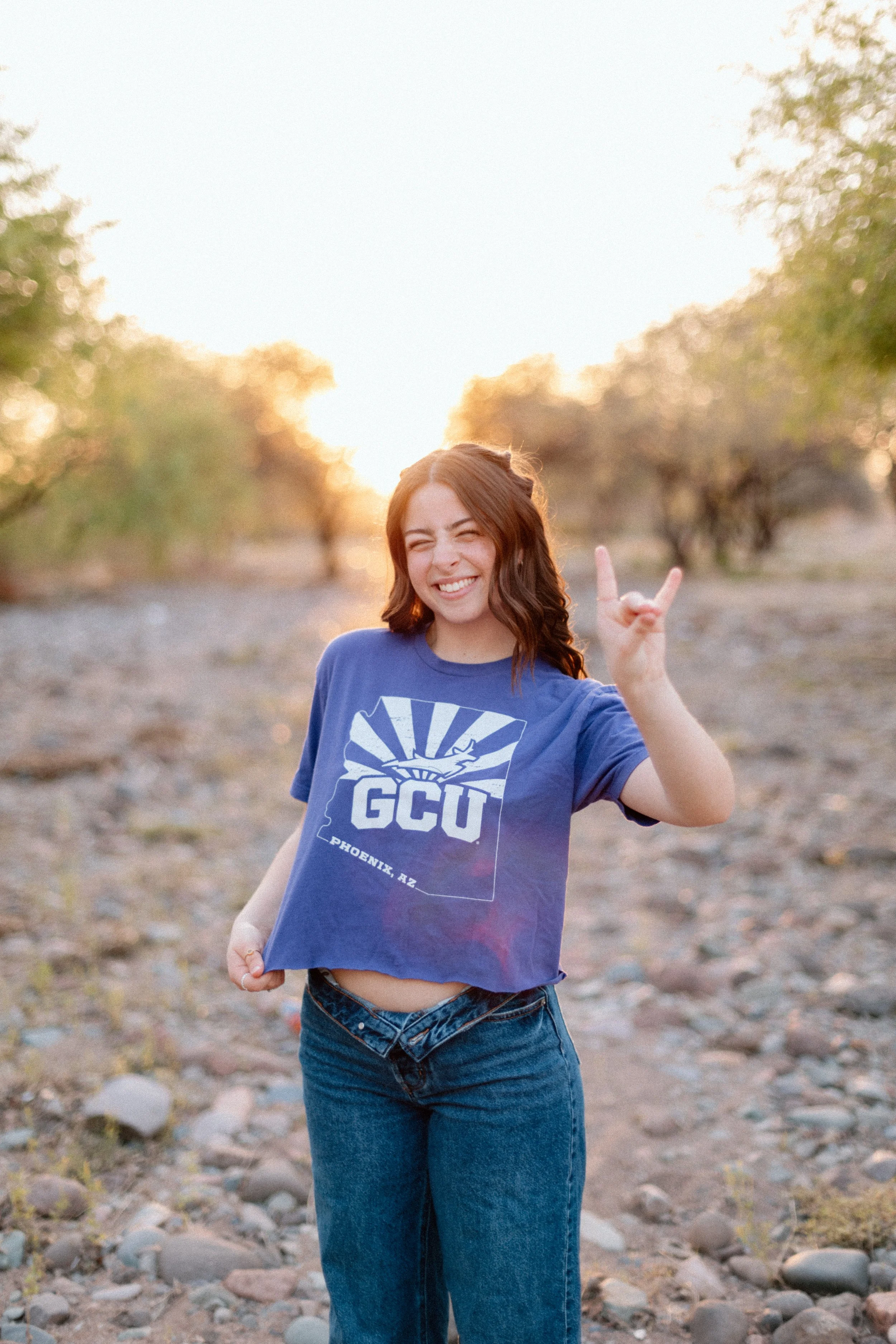 Young woman with dark brown hair smiling and making a 'rock on' gesture with her right hand, standing outdoors in a rocky area with trees and sunset in the background, wearing a purple GCU T-shirt and blue jeans.