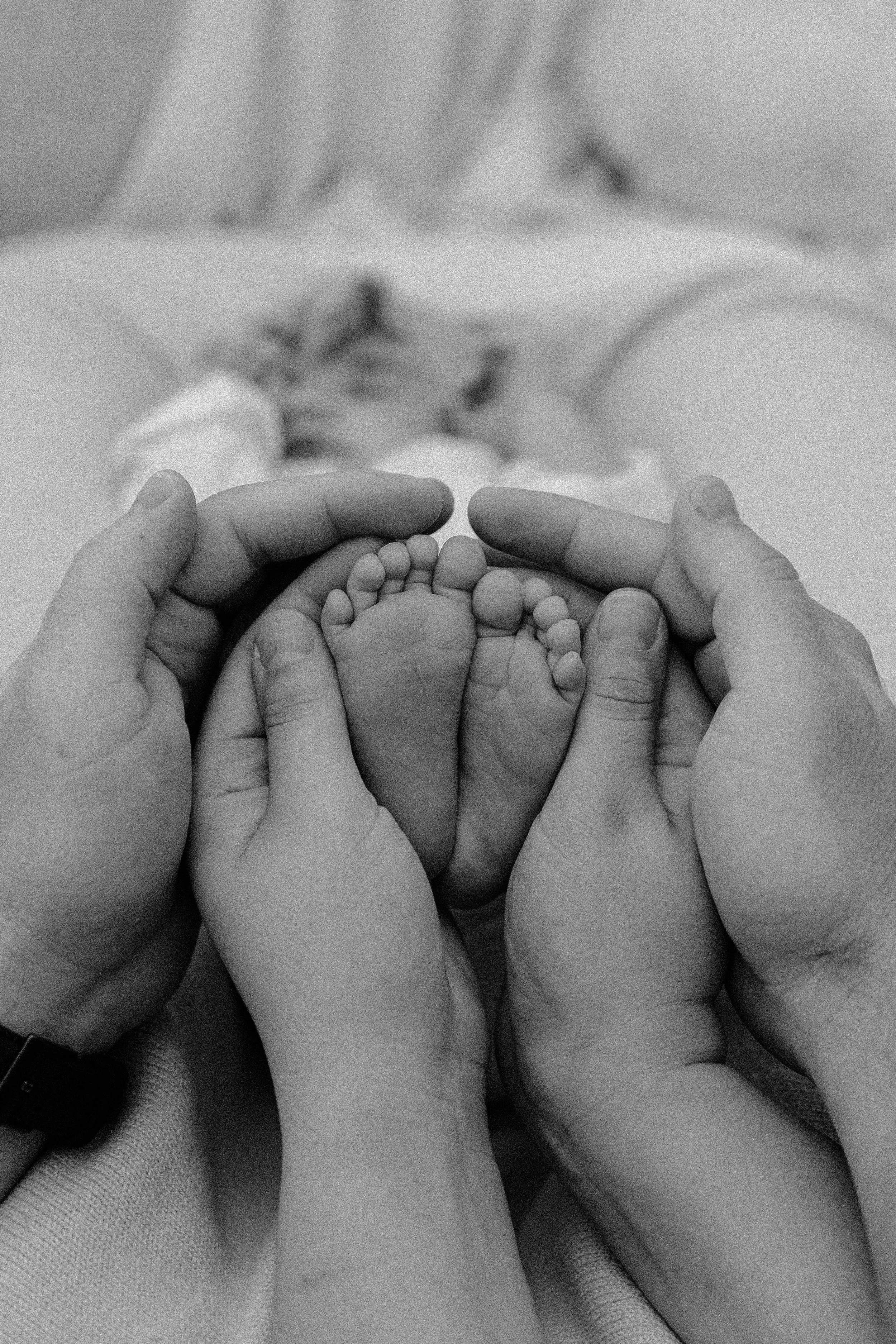 Close-up of a pair of adult hands holding tiny baby feet, with a blurred background of a baby lying in a hospital bed.