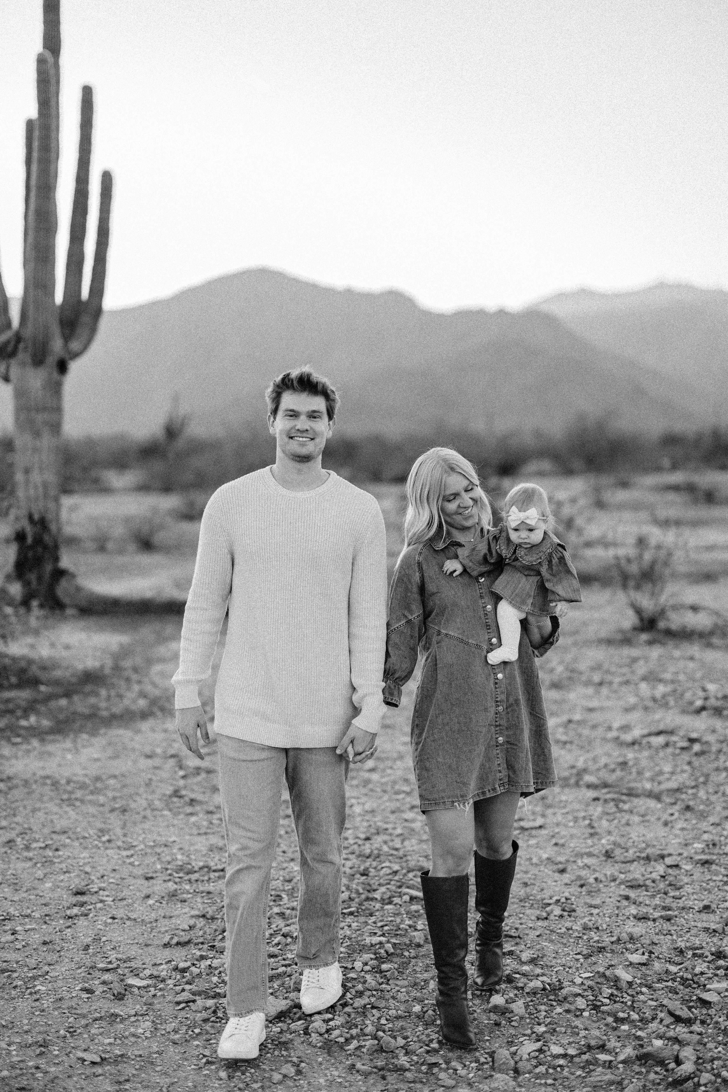 A family of three walking in a desert landscape with cacti and mountains in the background, black and white photo.