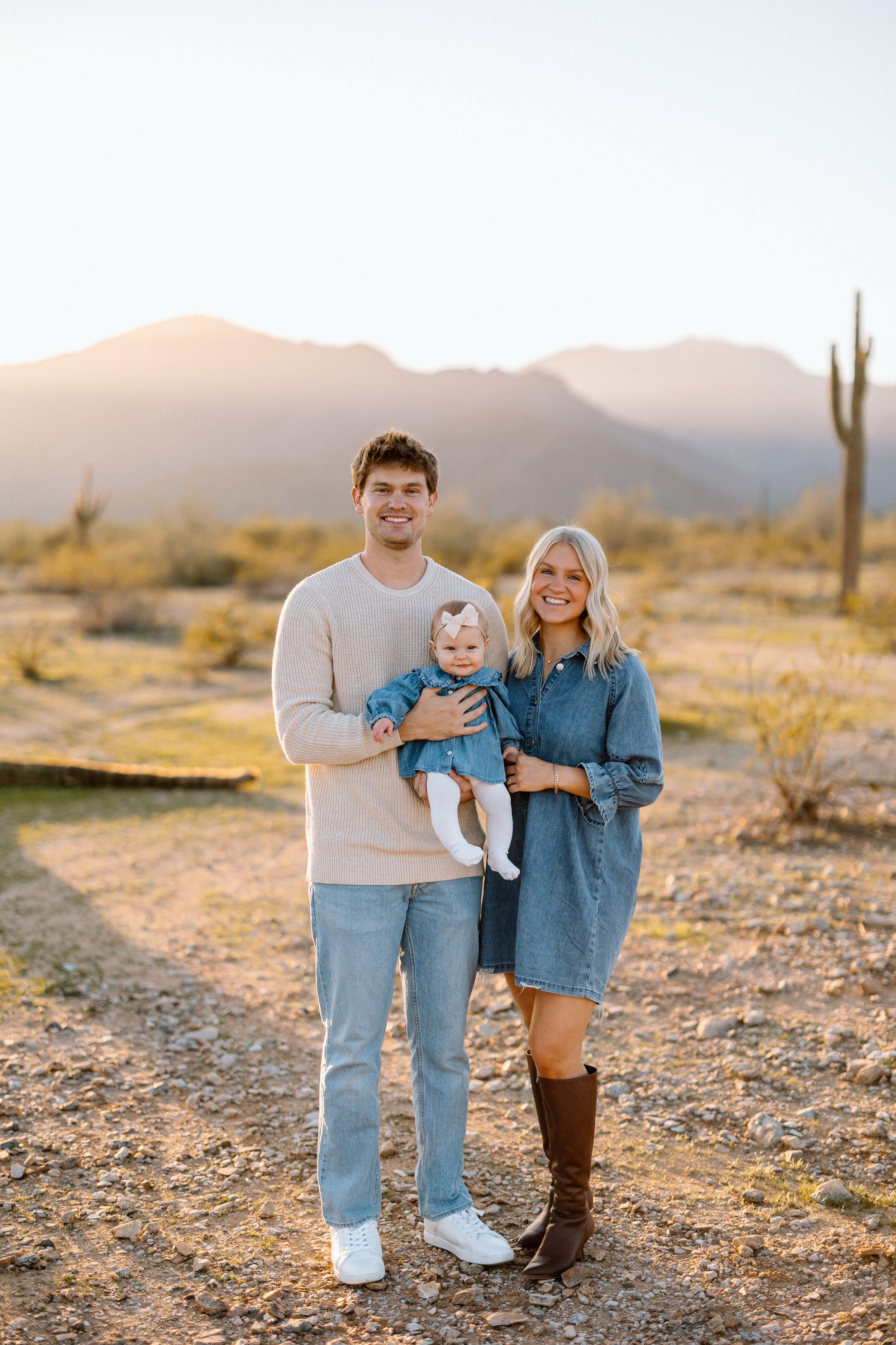 A family of three, a man, a woman, and a young girl, posing outdoors in a desert landscape with mountains and cacti in the background during sunset.