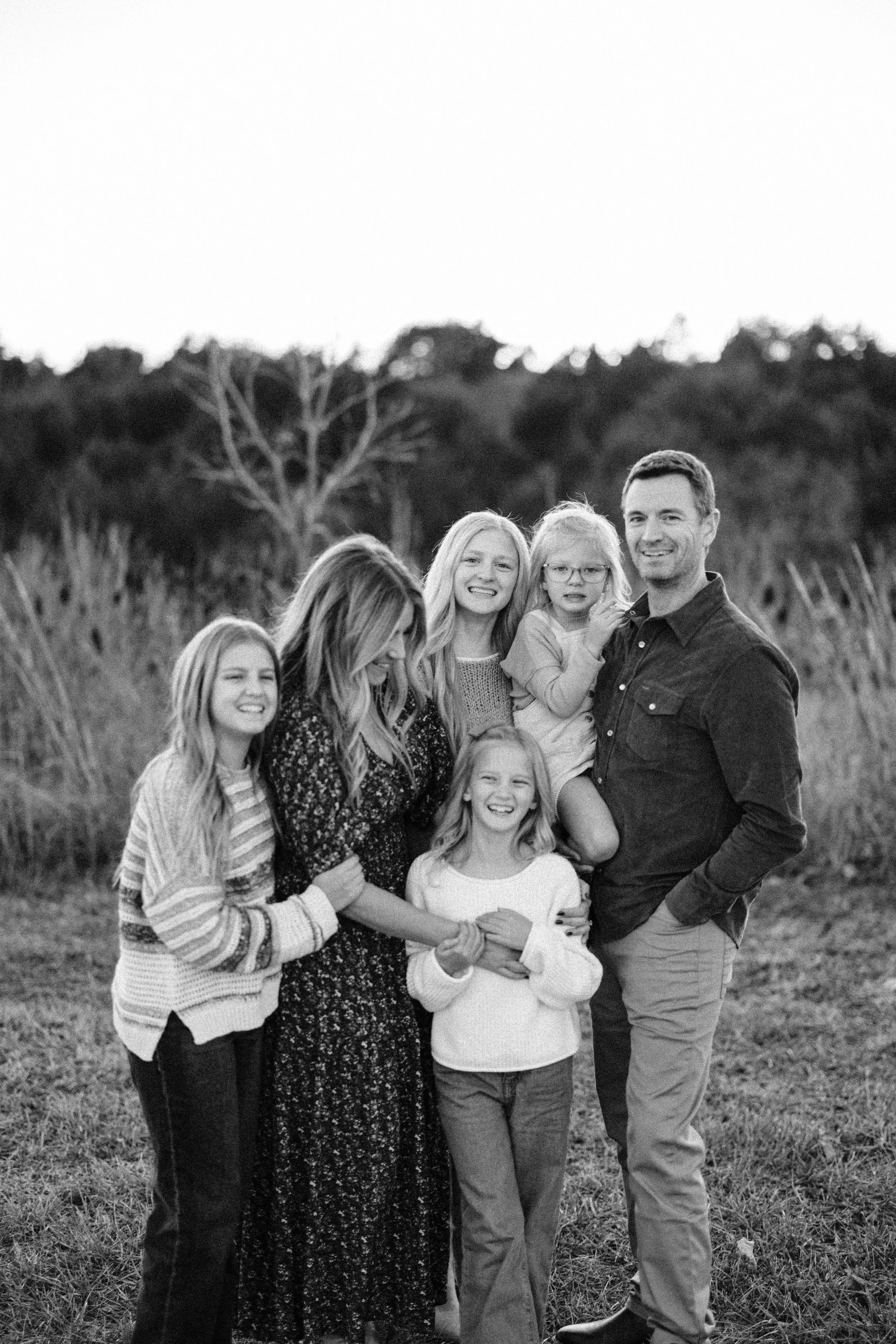 A black and white photo of a family of six standing outdoors on a grassy field, smiling and hugging each other, with trees and bushes in the background.