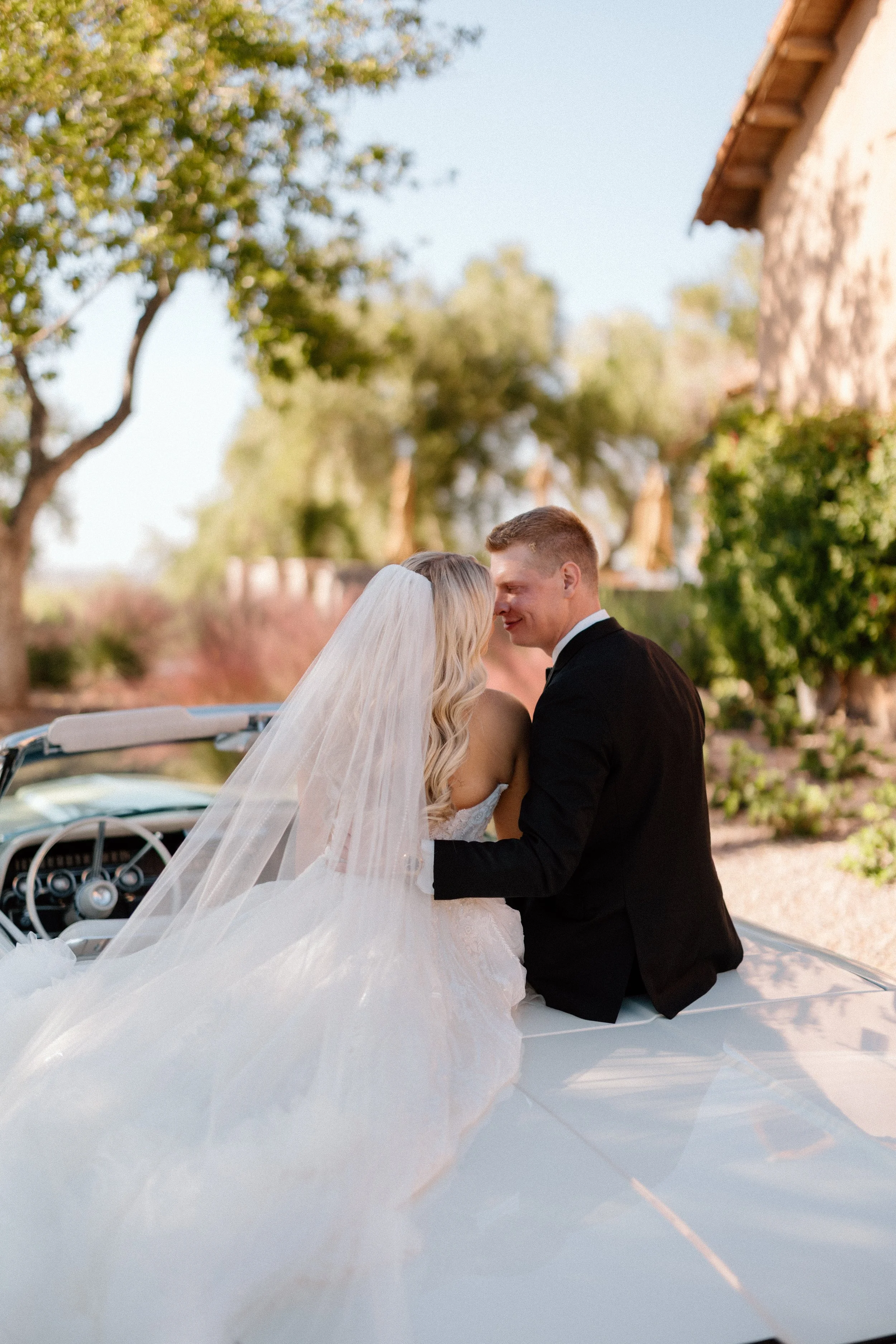 A bride and groom sitting on a vintage white car outdoors, close to each other, with the bride wearing a white wedding dress and veil and the groom in a black tuxedo, surrounded by trees and greenery.