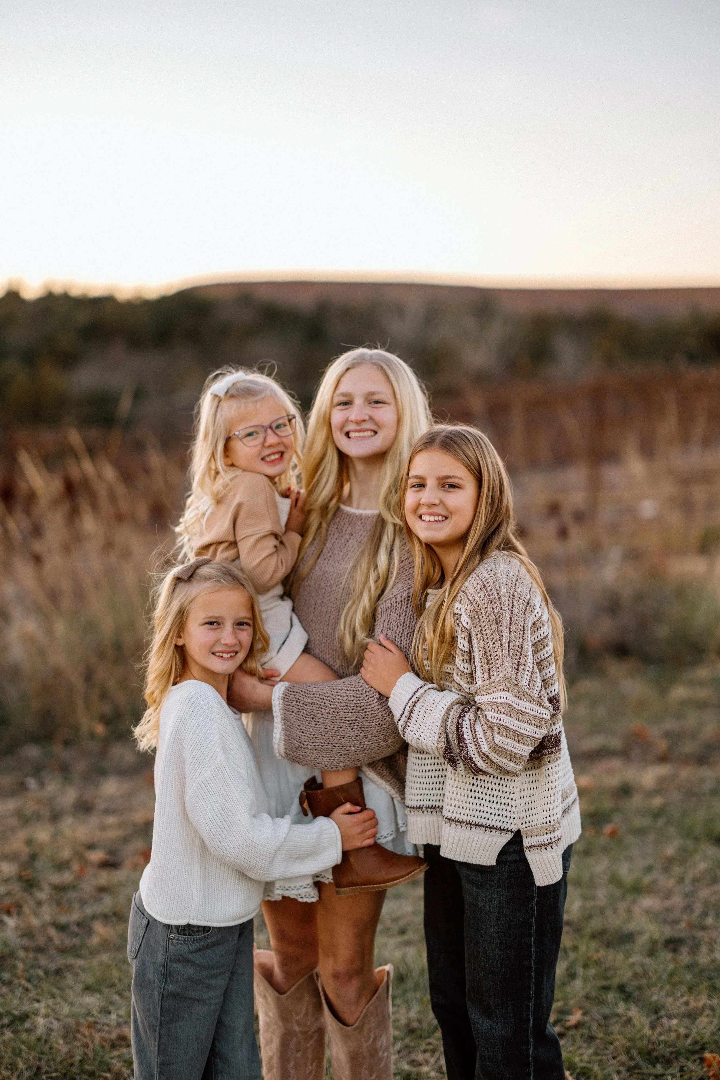 A woman with four young girls outdoors during sunset, all smiling and dressed in cozy winter clothing.