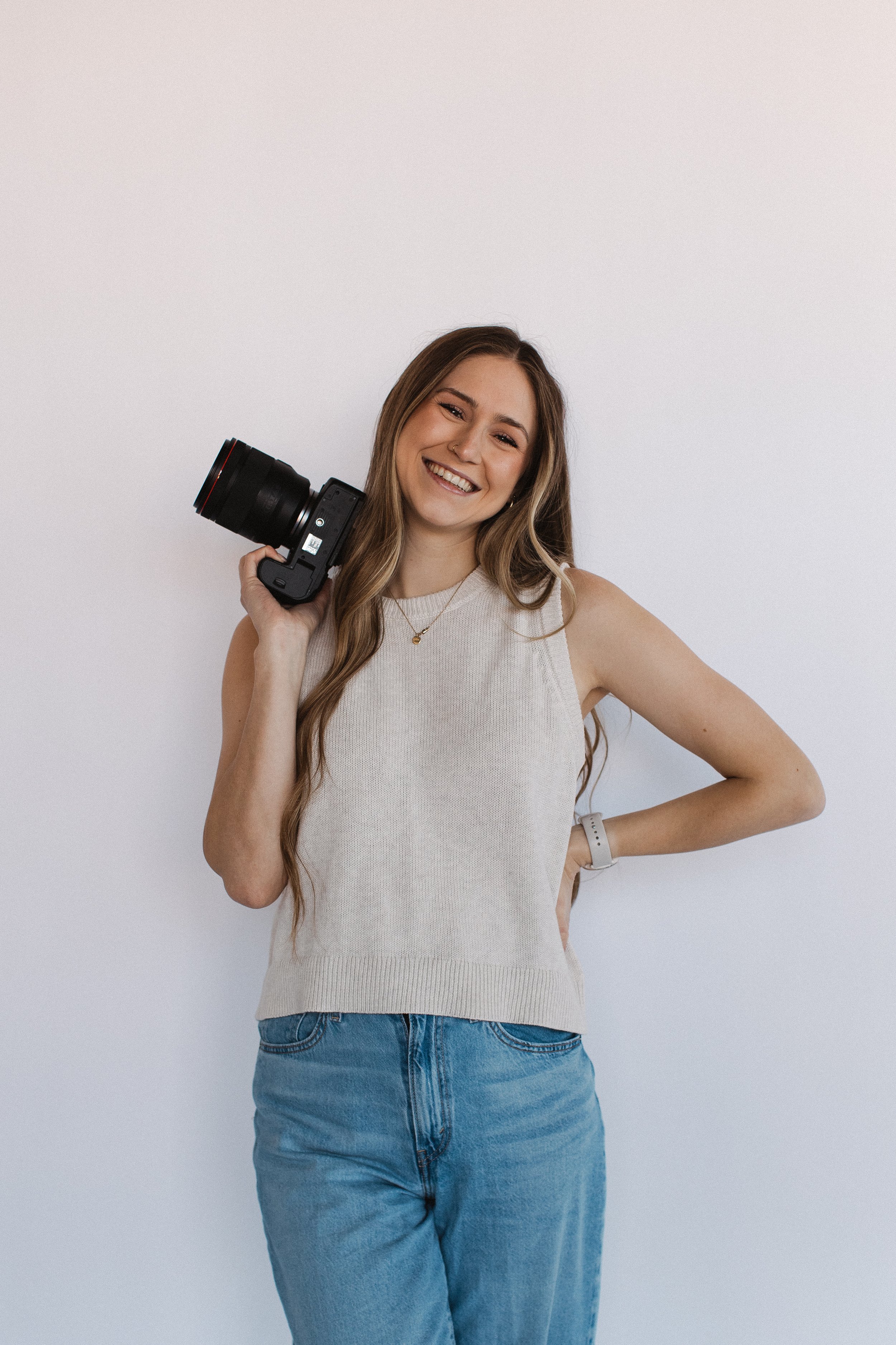 A young woman with long brown hair, smiling, holding a camera on her right shoulder, standing against a plain white wall.
