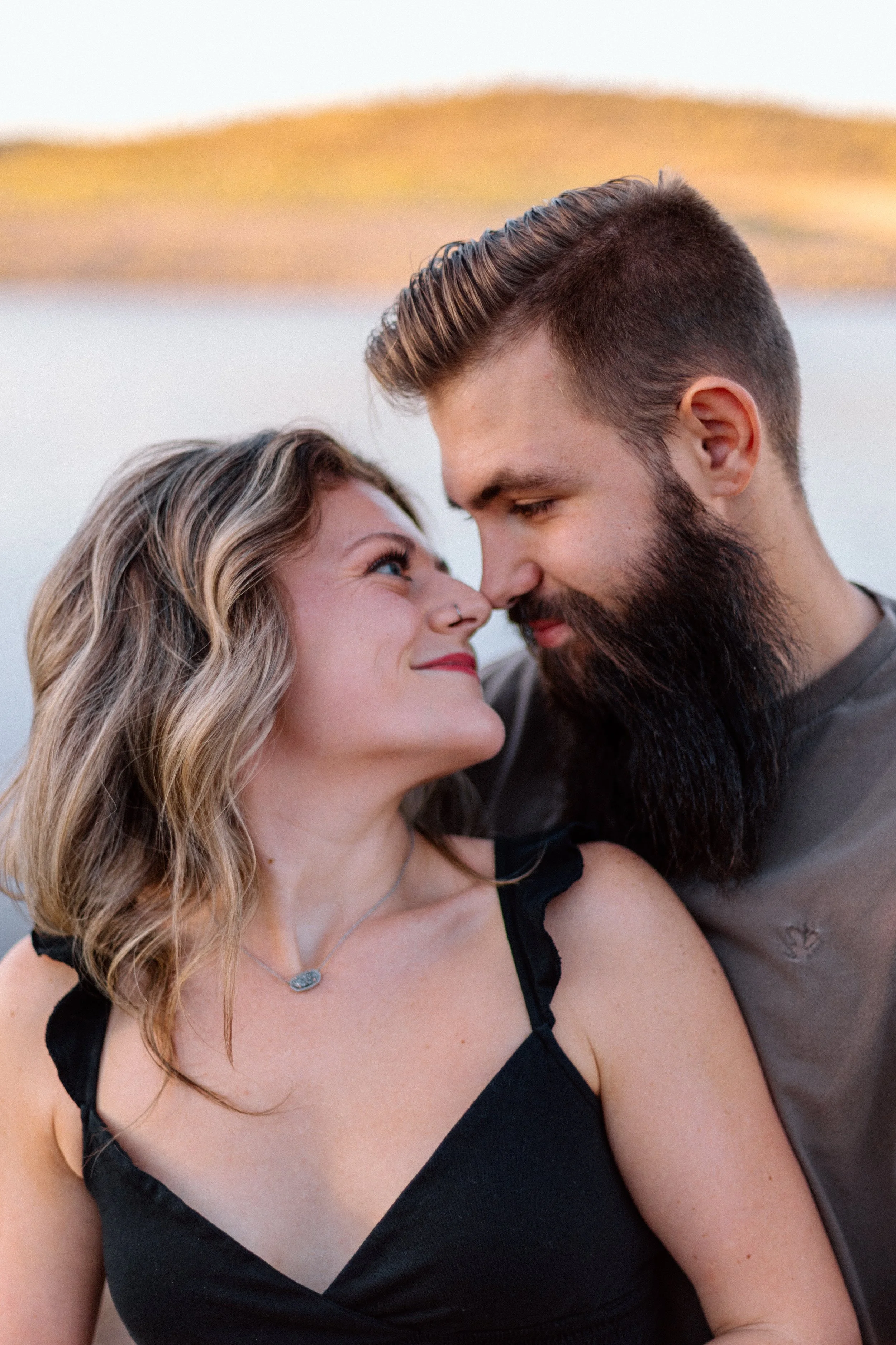 A couple standing close together outdoors, with hills and a body of water in the background, gazing into each other's eyes.