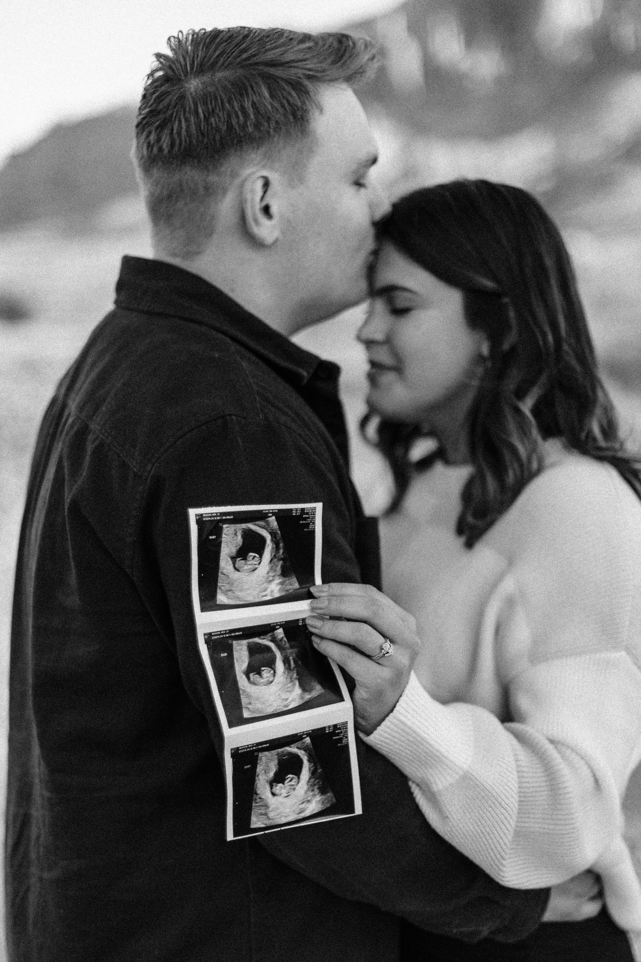 A black and white photo of a couple embracing, with the woman holding ultrasound pictures showing a fetus, indicating an announcement of pregnancy.