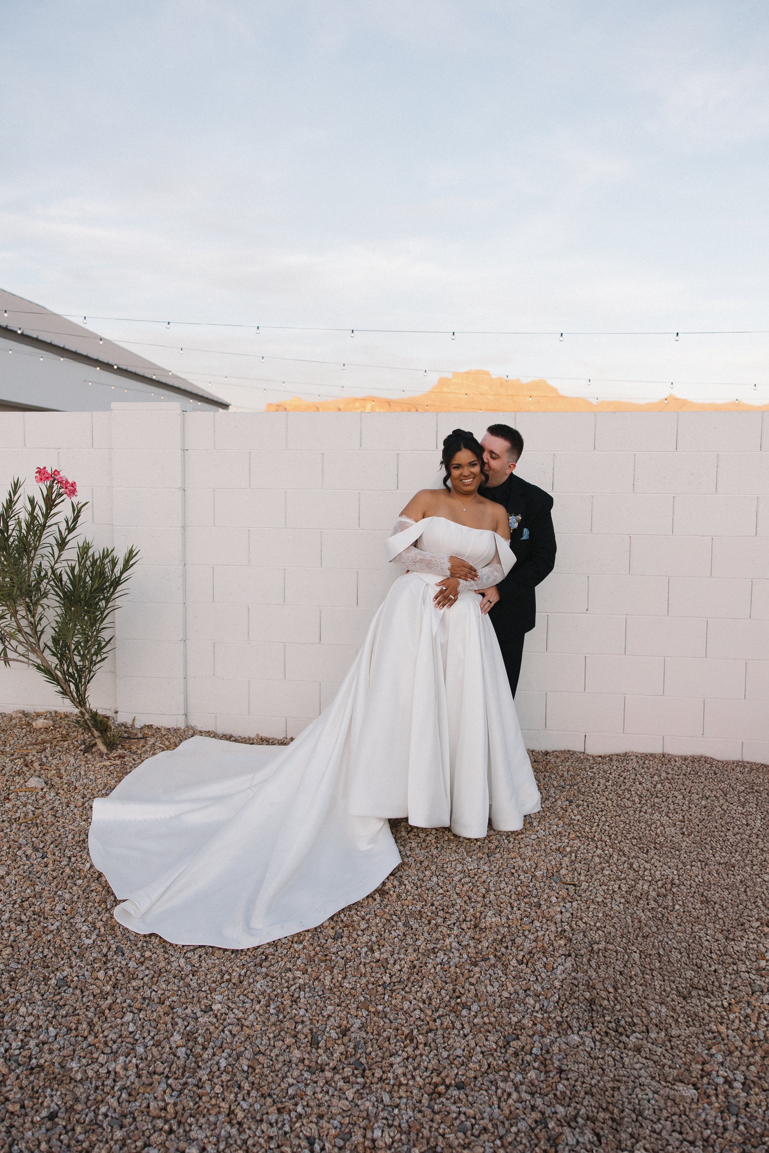Bride and groom hugging outdoors near a white brick wall, with desert mountains in the background and string lights overhead.