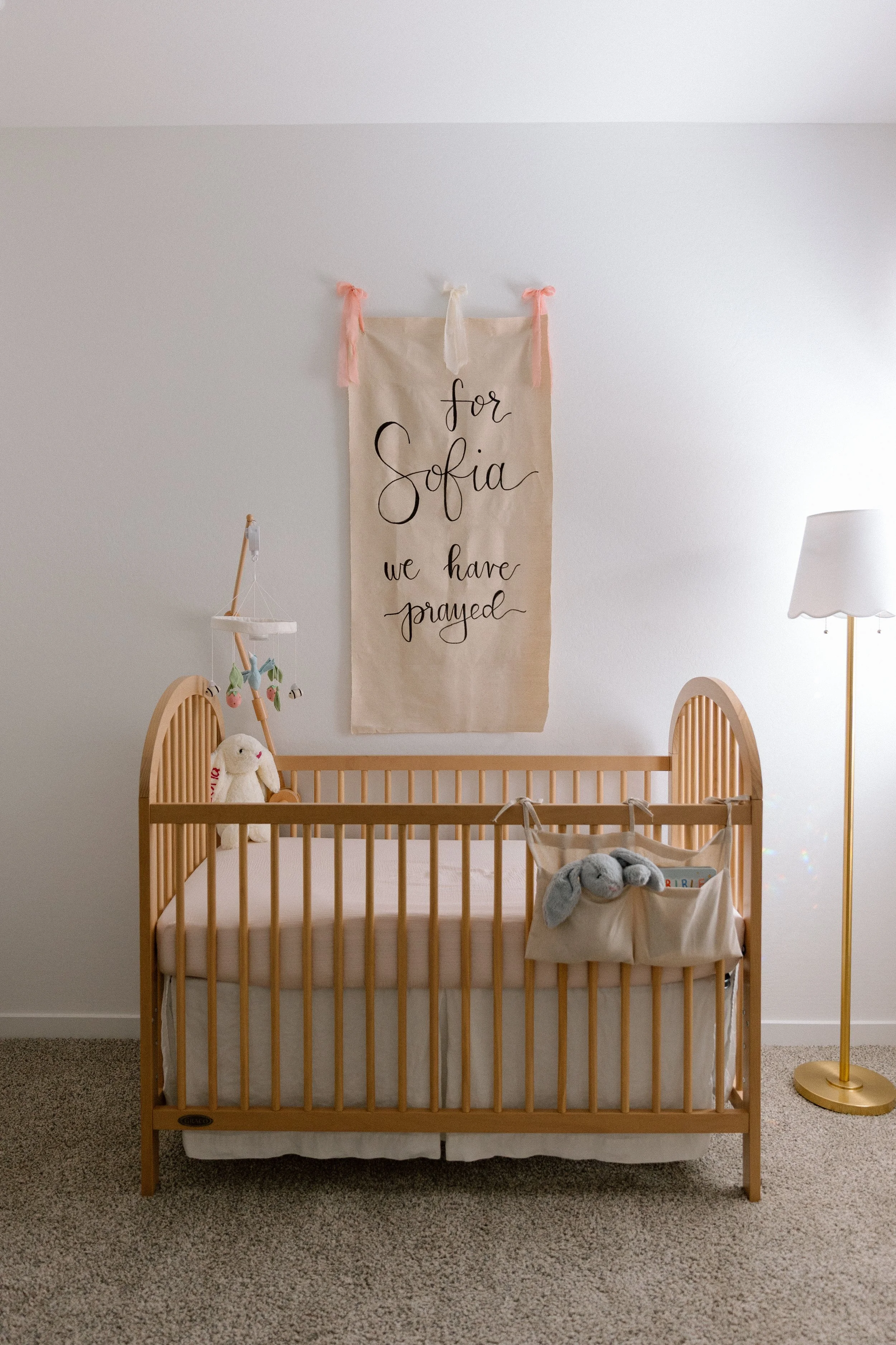 A nursery room with a wooden crib, stuffed animals, a hanging mobile, and a banner on the wall that says 'for Sofia we have prayed'.