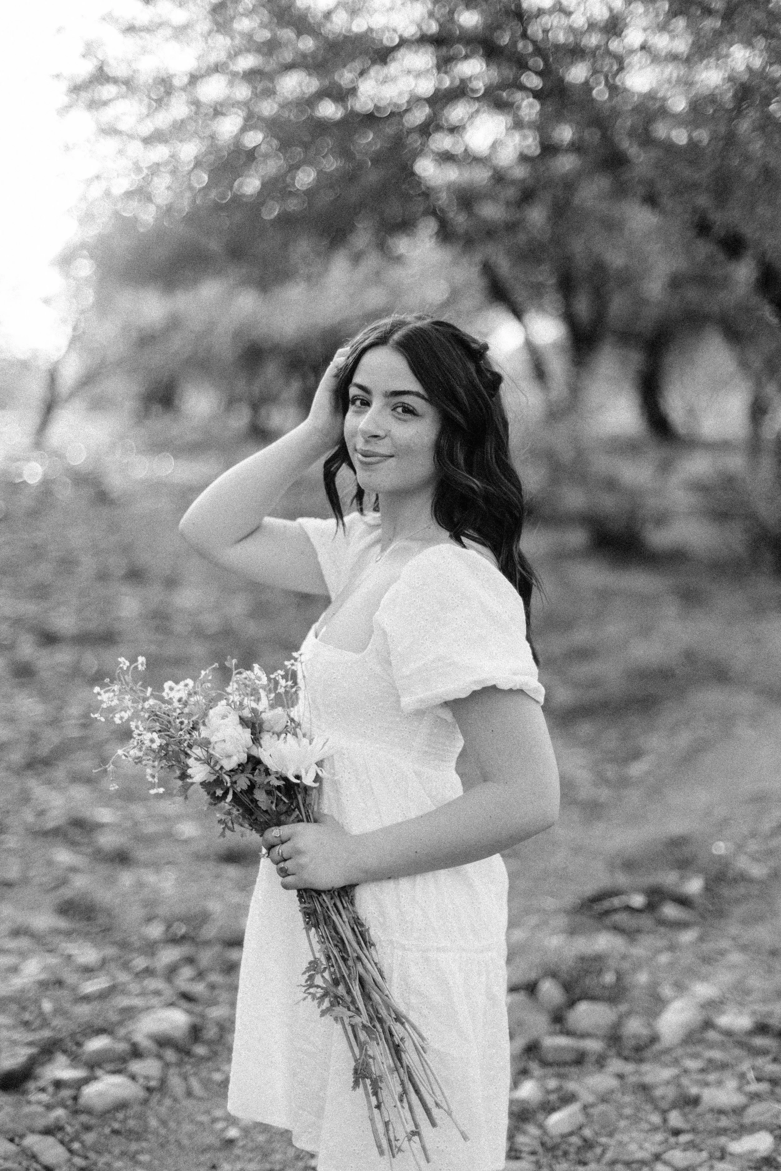 A woman in a white dress standing outdoors on rocky ground, holding a bouquet of flowers, with trees in the background, smiling and touching her hair.
