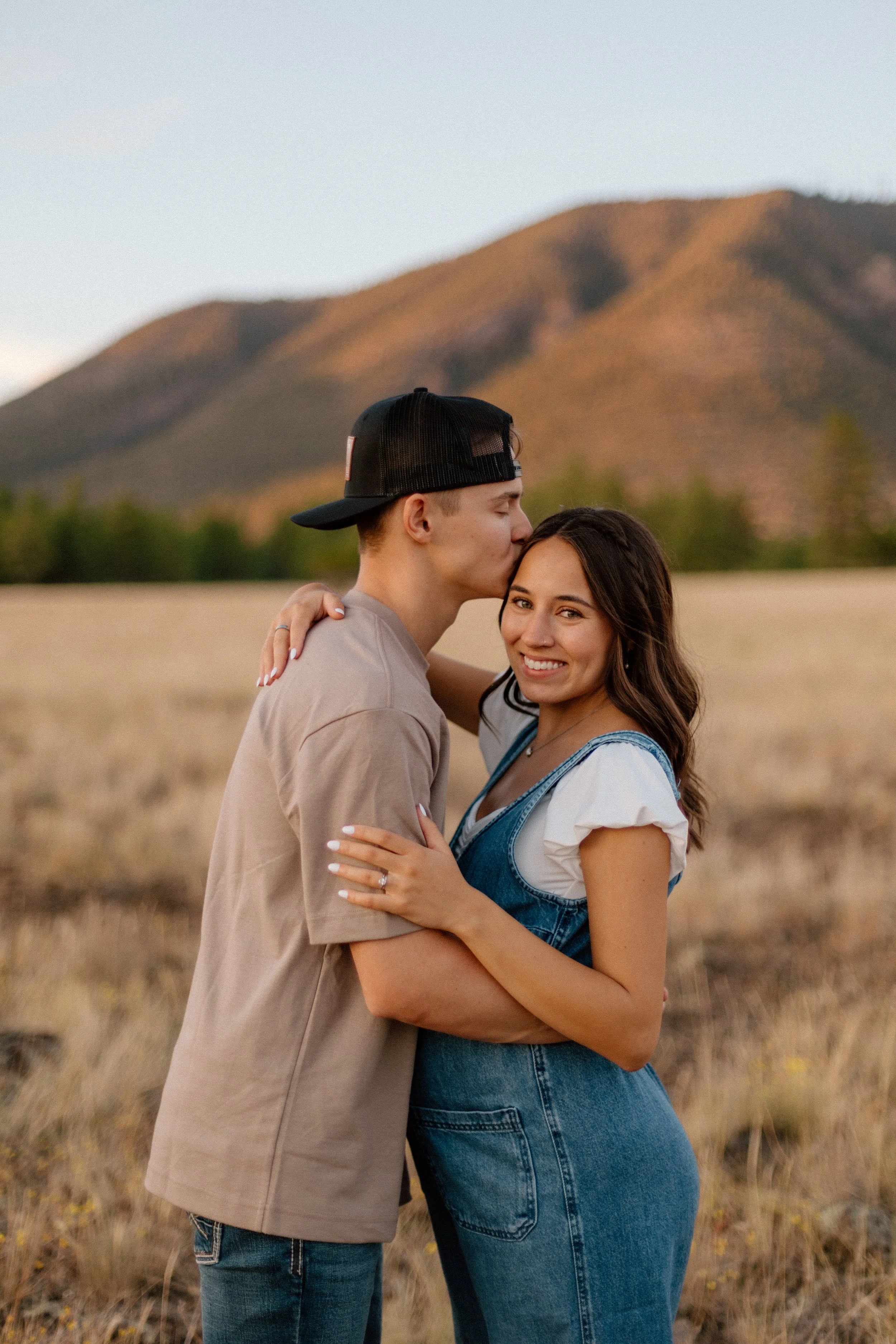 A young couple embracing and smiling in a grassy field with mountains in the background during sunset, the man is kissing the woman's forehead.