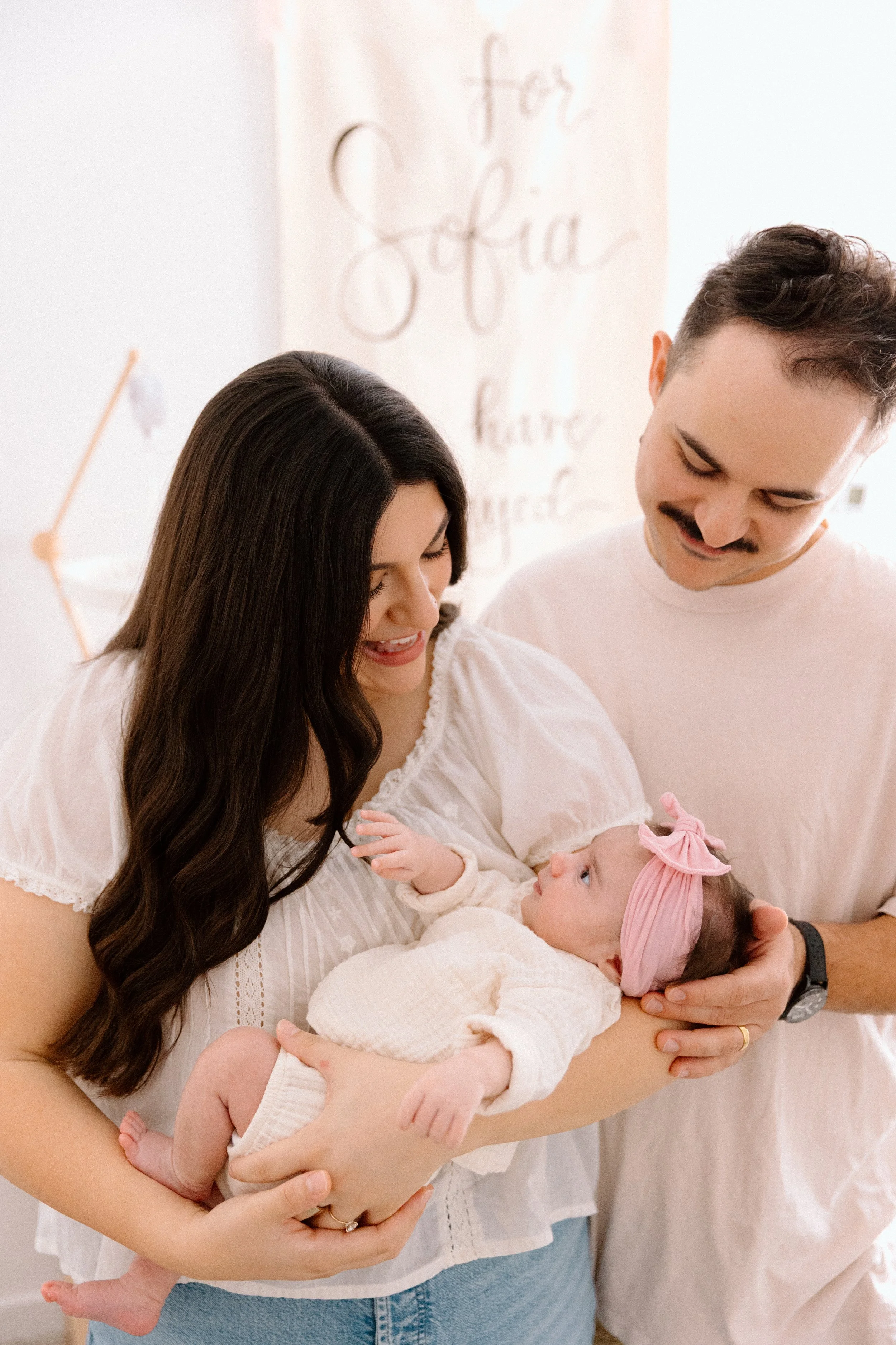 A woman holding a newborn baby with a man standing beside her, all smiling in a nursery decorated with soft colors and a wall hanging that reads 'for Sofia'.