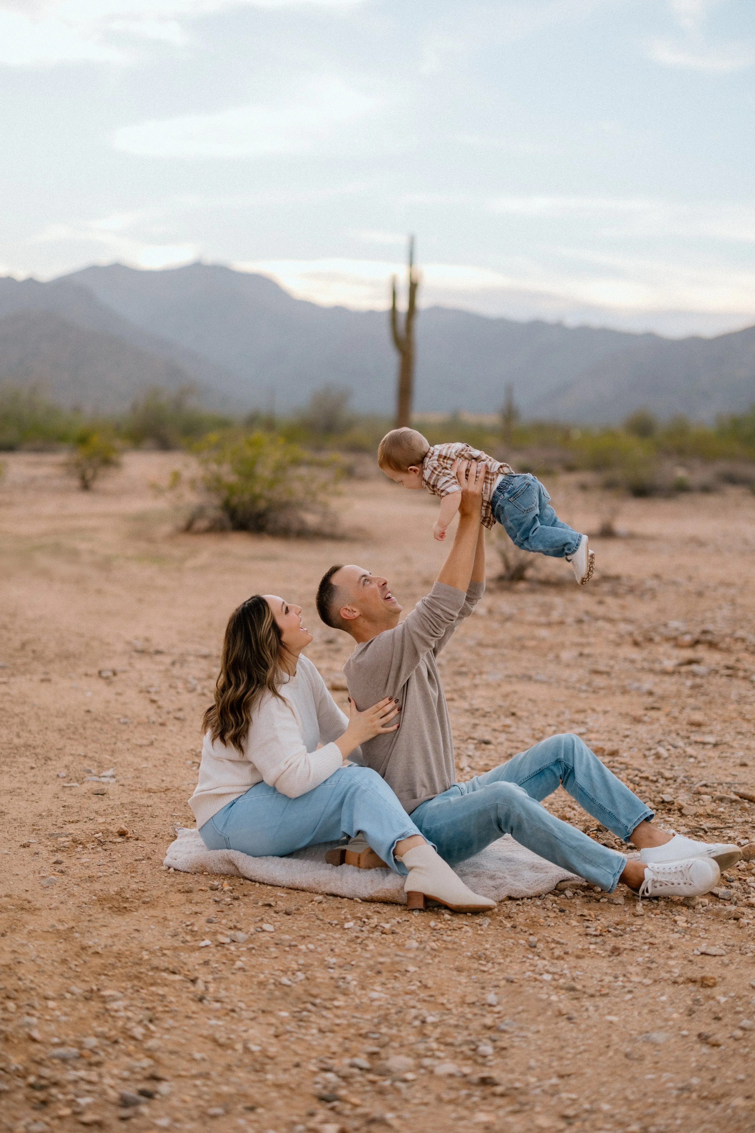 A family of three enjoying time outdoors in a desert landscape, with mountains in the background. The father is sitting on a blanket, lifting their young son into the air, while the mother sits beside them, smiling and watching.