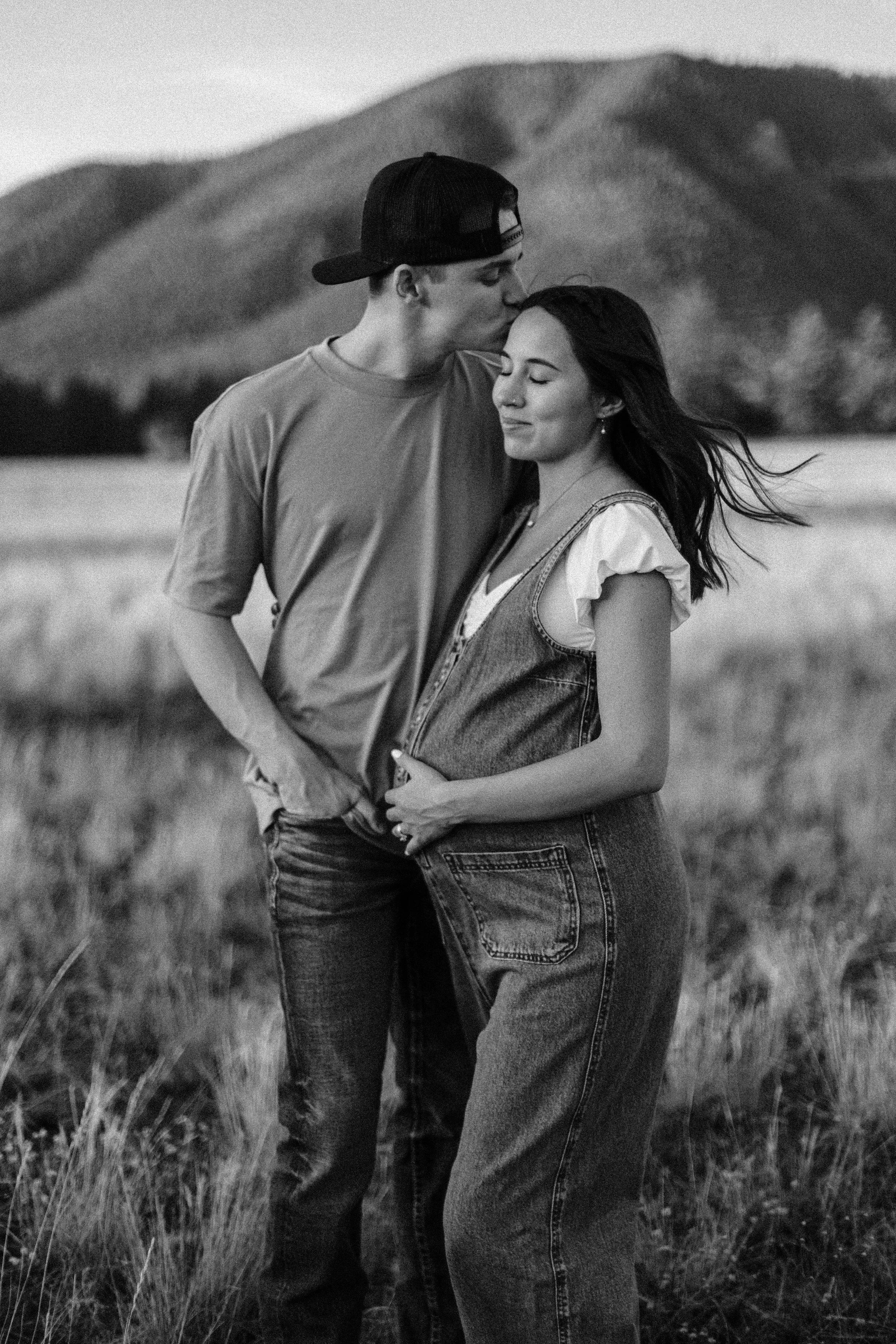 A young couple standing in a field with mountains in the background, gently embracing and sharing a tender moment, black and white photo