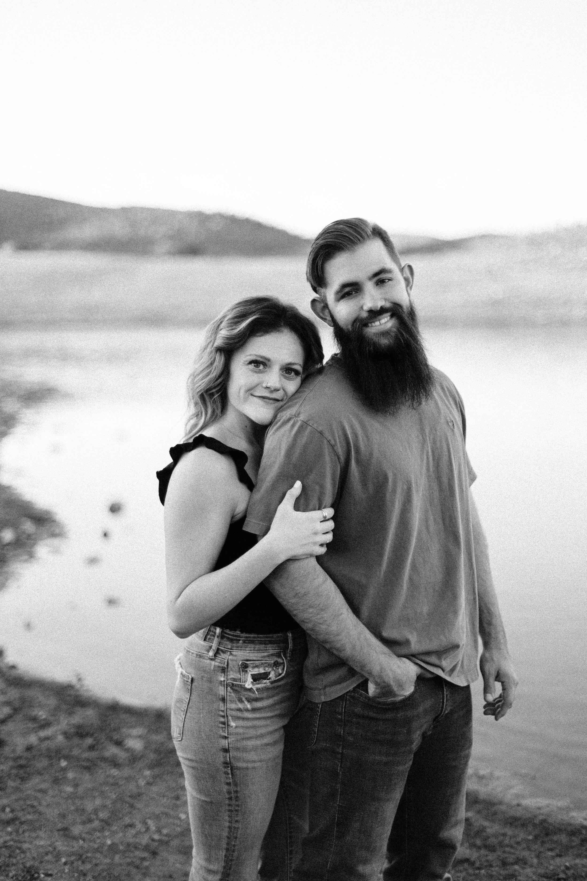 A black and white photo of a smiling couple standing outdoors near a body of water, with hills in the background.