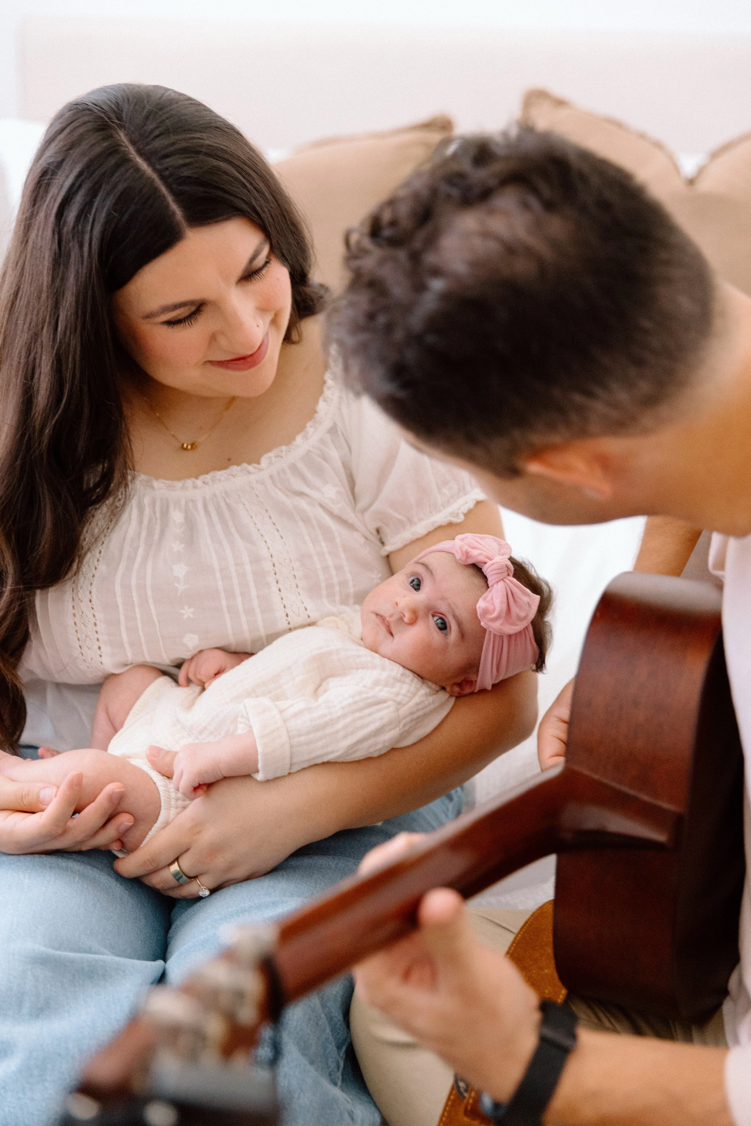 A woman holding a baby girl with a pink headband, while a man plays guitar and sings to them.