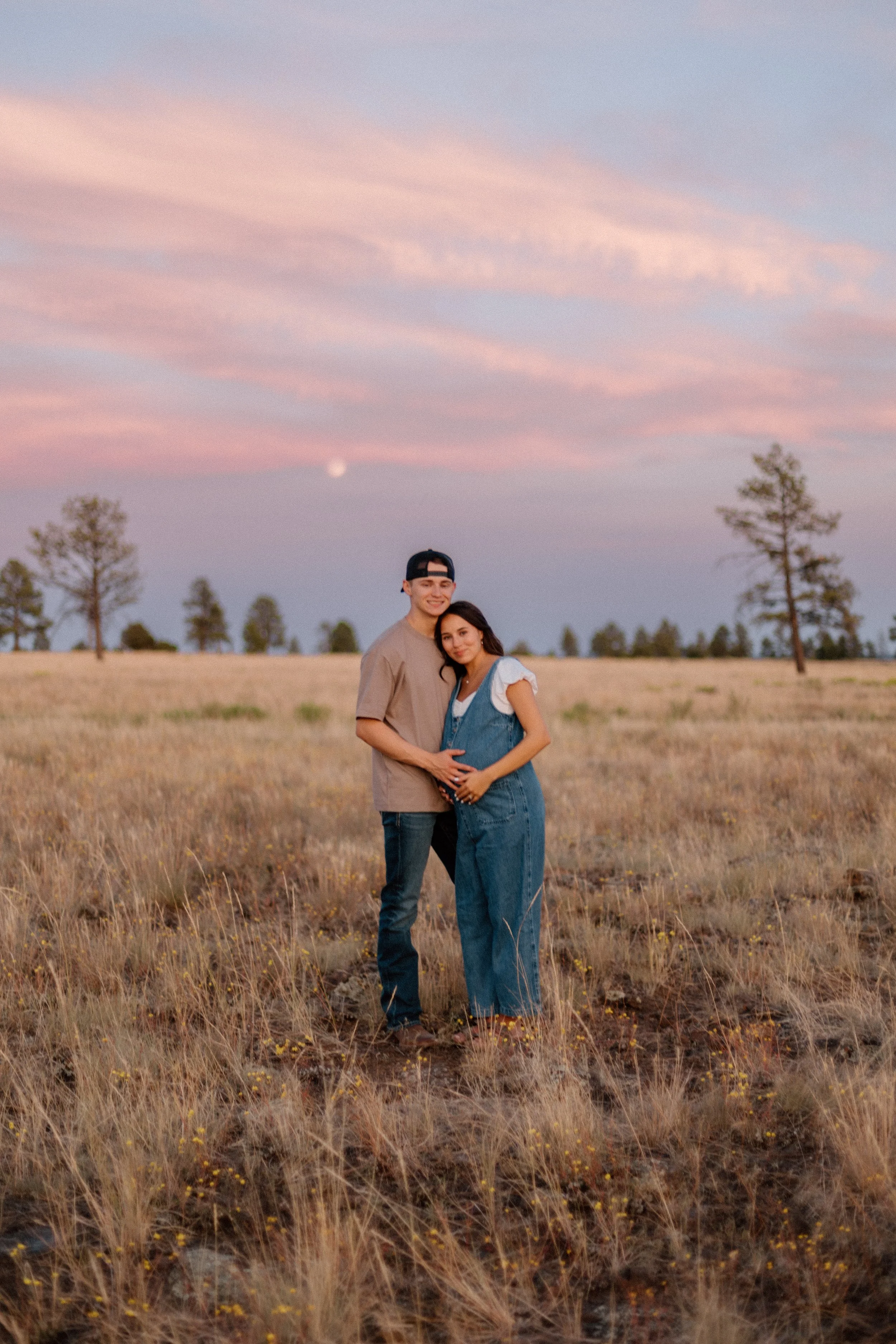 A young couple stands together in a grassy field during sunset, with the woman pregnant and the man embracing her belly, smiling and looking happy.