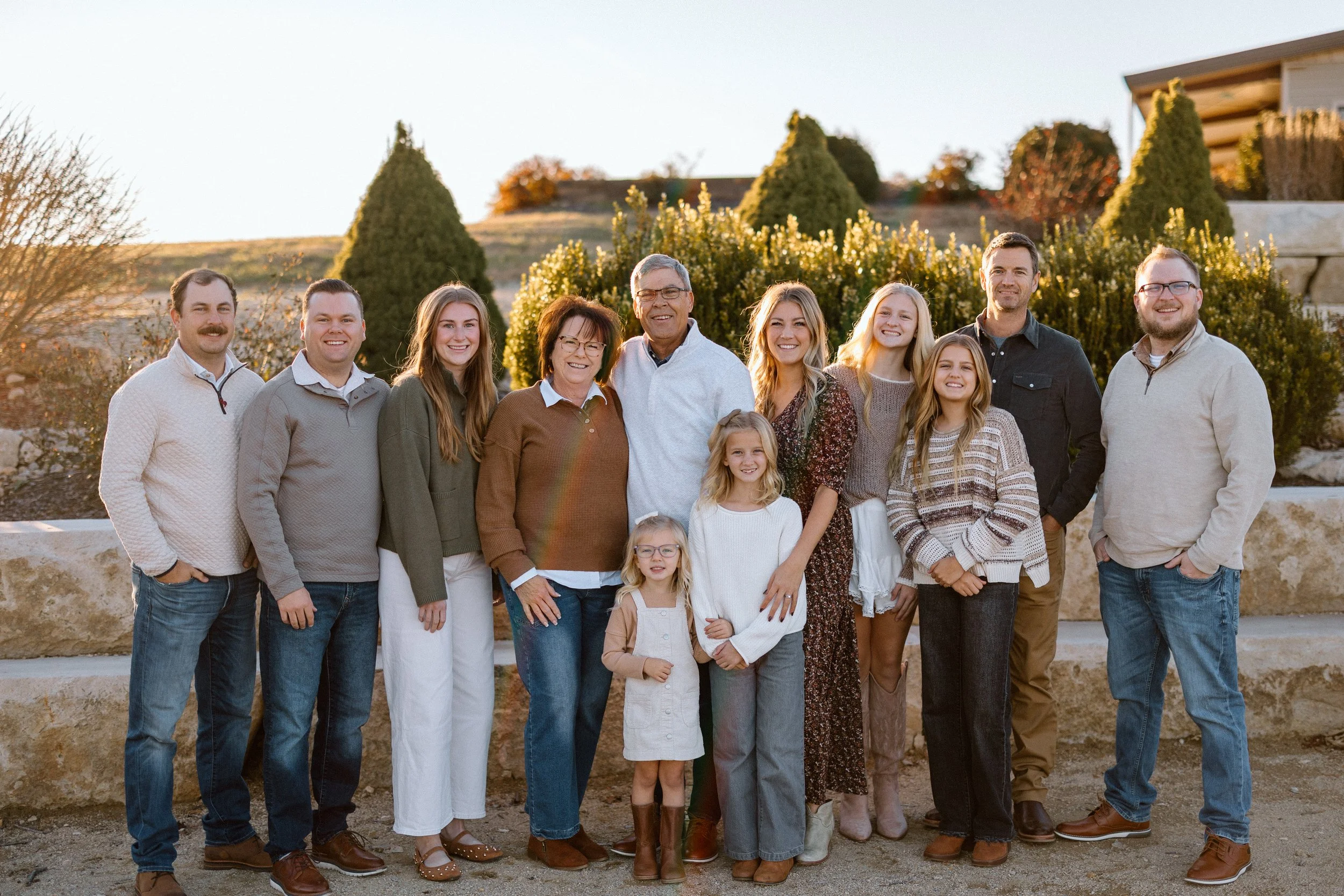 Family group photo outdoors during golden hour, with greenery and rocks in the background, featuring adults and children smiling.