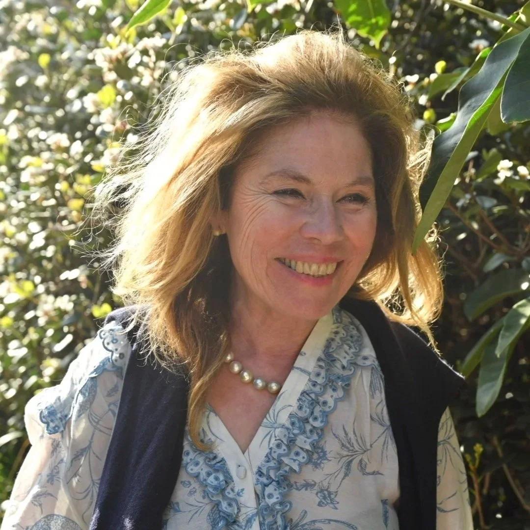 A smiling female naturopath outdoors in sunlight, surrounded by green foliage.