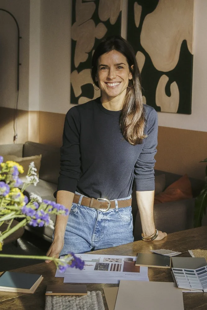 A woman with dark hair, smiling, standing indoors at a table with design samples and a bouquet of purple flowers.