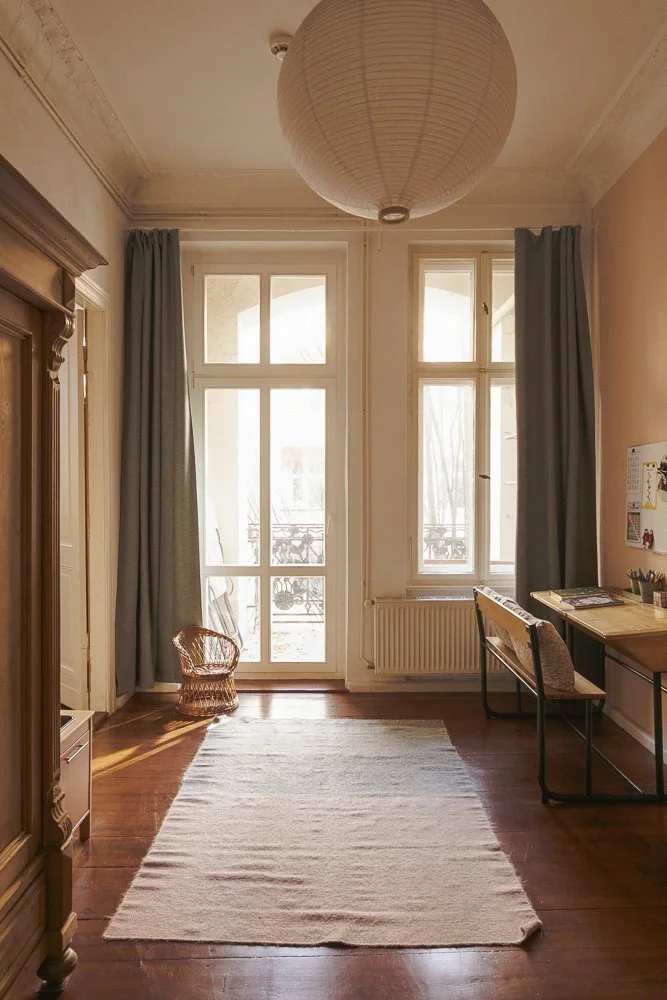 Bright room with wooden floor, large windows and doors leading outside, curtains, a small wicker chair, a rug, a desk with supplies, and a hanging paper lantern ceiling light.