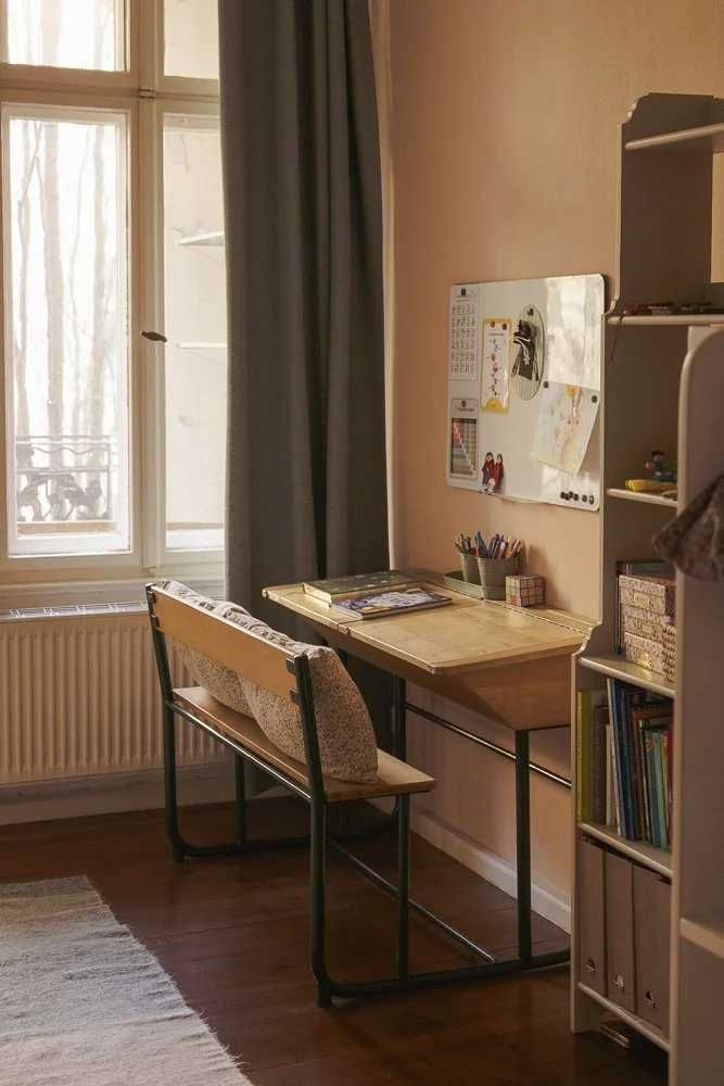 A small study corner with a wooden desk, a wooden bench with patterned cushions, a white bookcase, and a whiteboard on the wall. Sunlight comes through a window with closed dark curtains.