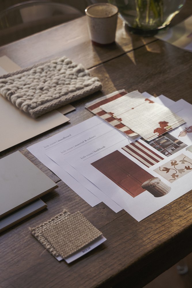 A wooden desk with fabric swatches, paint samples, and design plans, including a knitted beige fabric, candle, and papers with interior decorating ideas.
