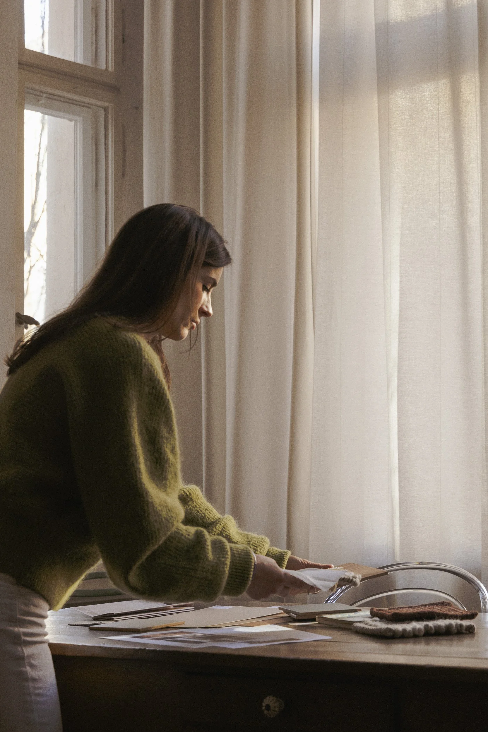 Woman organizing papers on a wooden table near a window with white curtains, sunlight streaming in.
