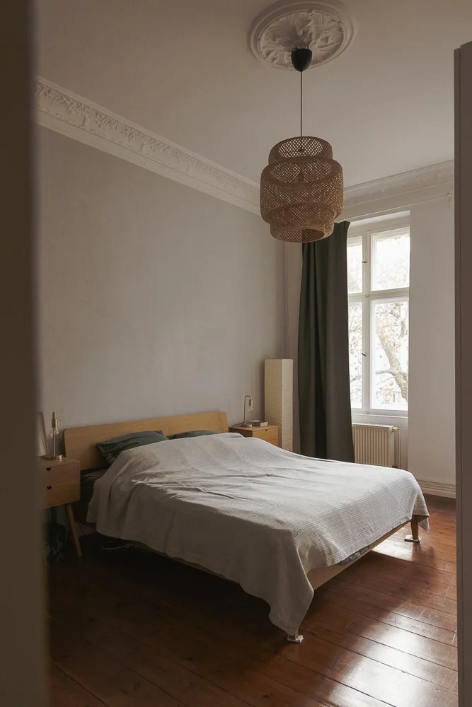 A minimalist bedroom with a wooden bed, white bedding, dark pillows, a wooden nightstand, a tall beige floor lamp, and a large window with dark curtains, in a room with white walls and hardwood floors, illuminated by natural light.