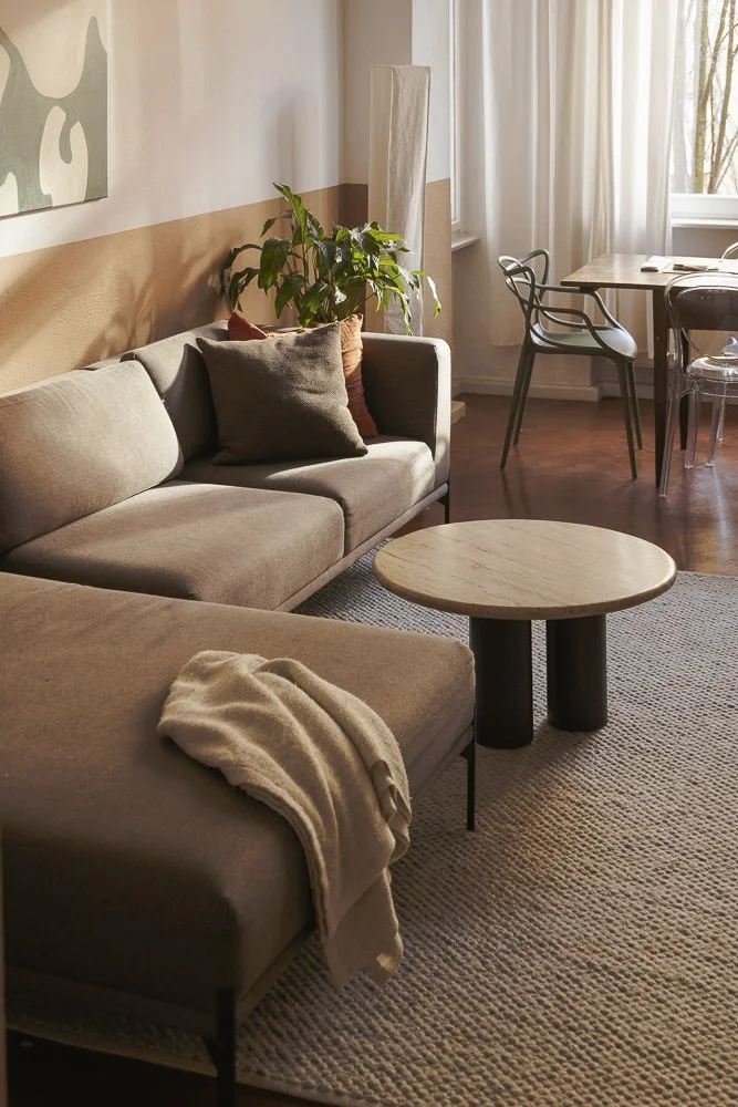 Living room with taupe sofa and throw blanket, round coffee table, plants, and dining area with chairs near a window.