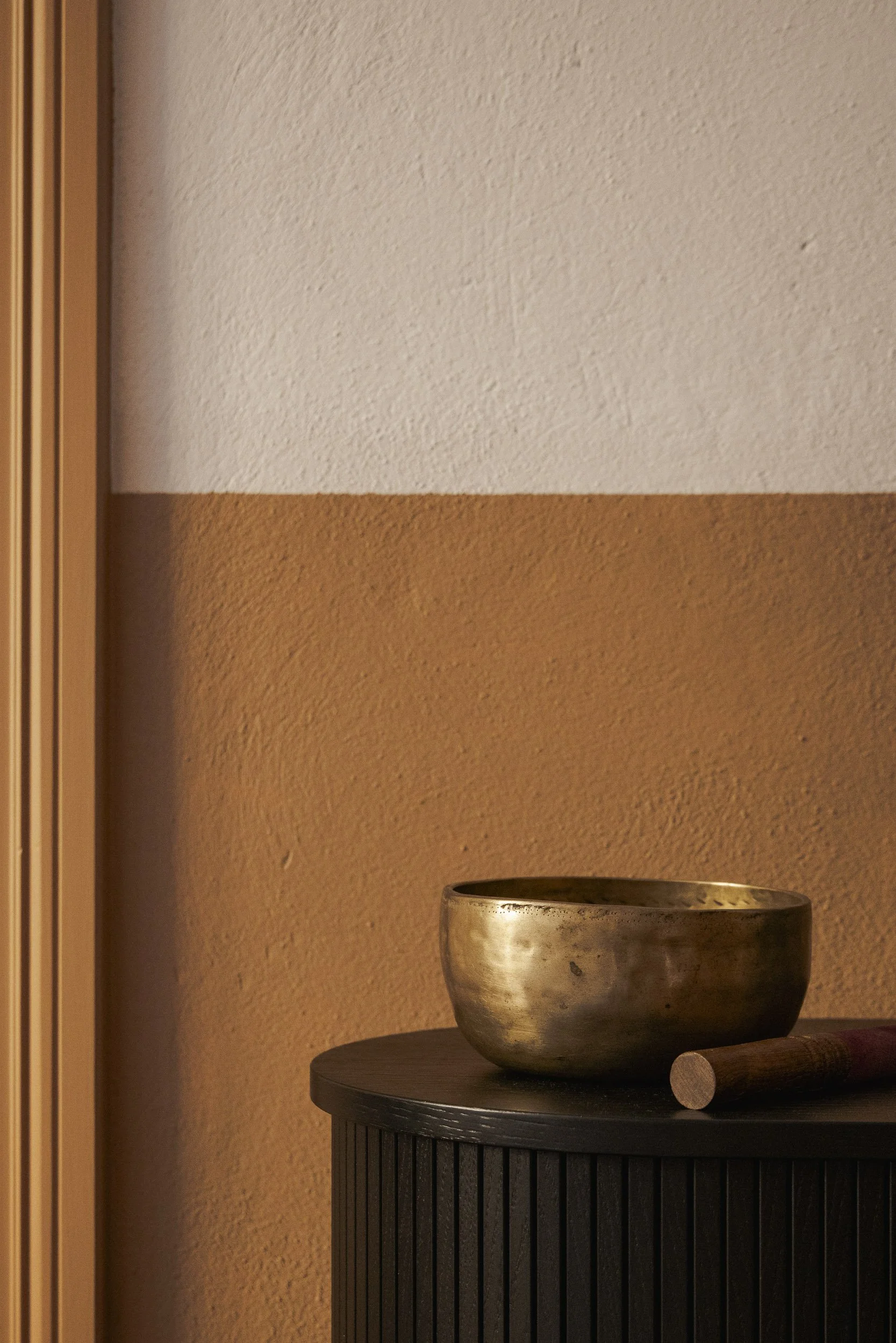 A metallic singing bowl and wooden mallet resting on a dark, rounded table against a two-tone wall, with the upper part painted white and the lower part painted brown.