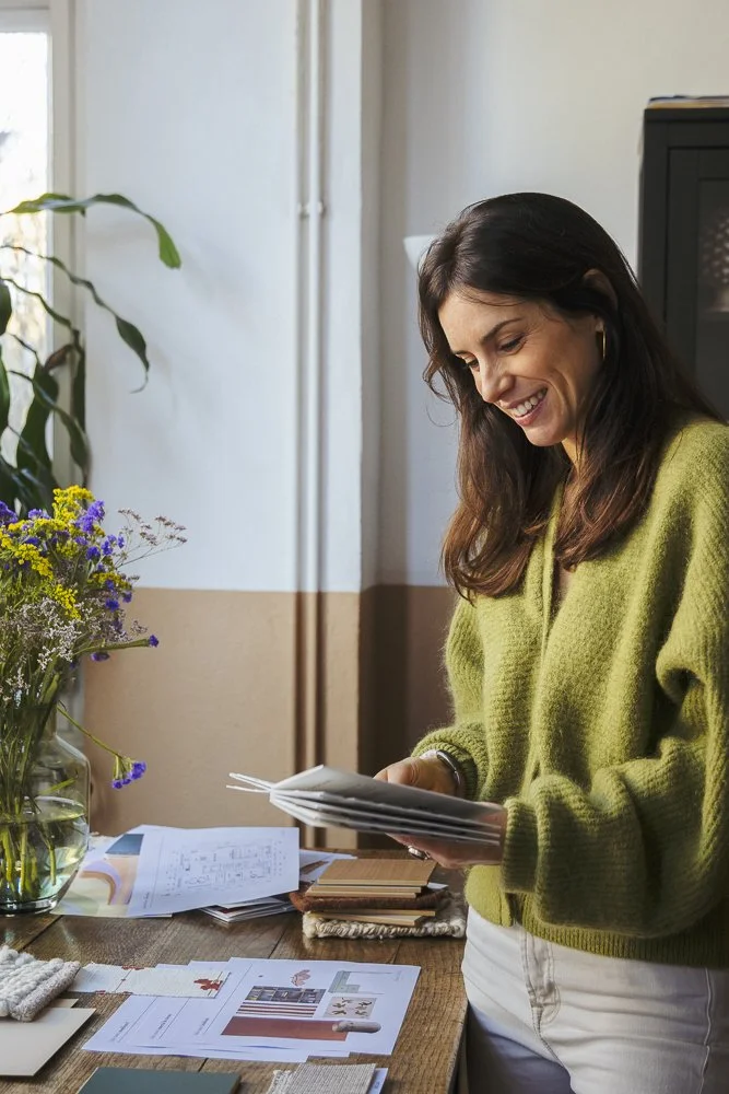 Woman with long brown hair wearing a green sweater looking at papers on a wooden table with various documents, books, and a vase of colorful flowers inside a bright room.