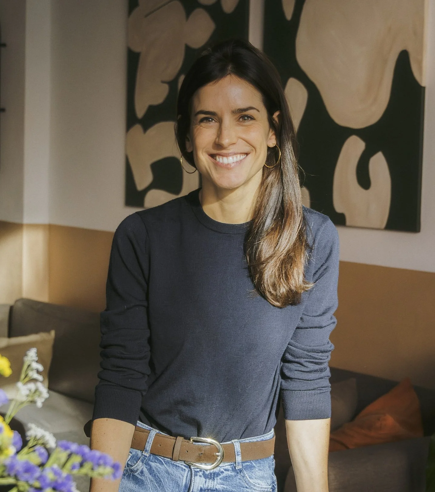A smiling woman with dark brown hair, wearing a dark blue sweater and light blue jeans, standing indoors with a floral painting in the background.