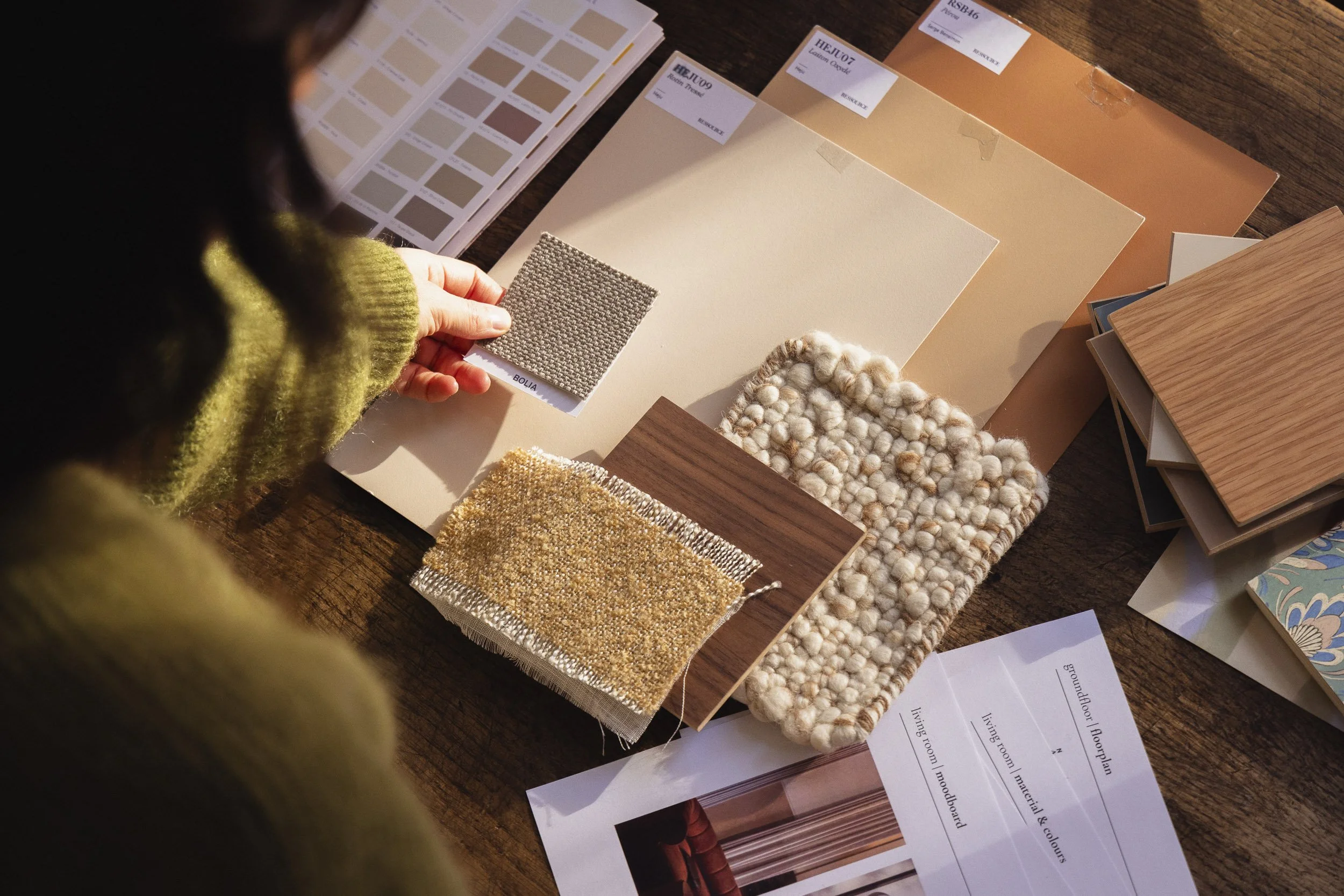 A person examining various fabric and material samples, including a color palette, fabric swatches, and wallpaper samples, on a wooden table.