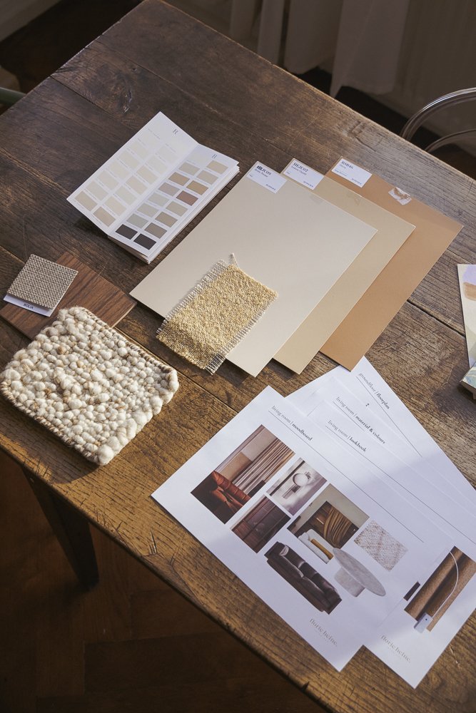 A wooden table with a color swatch booklet, fabric samples, paint chips, and interior design images on sheets of paper used for decorating or remodeling a room.