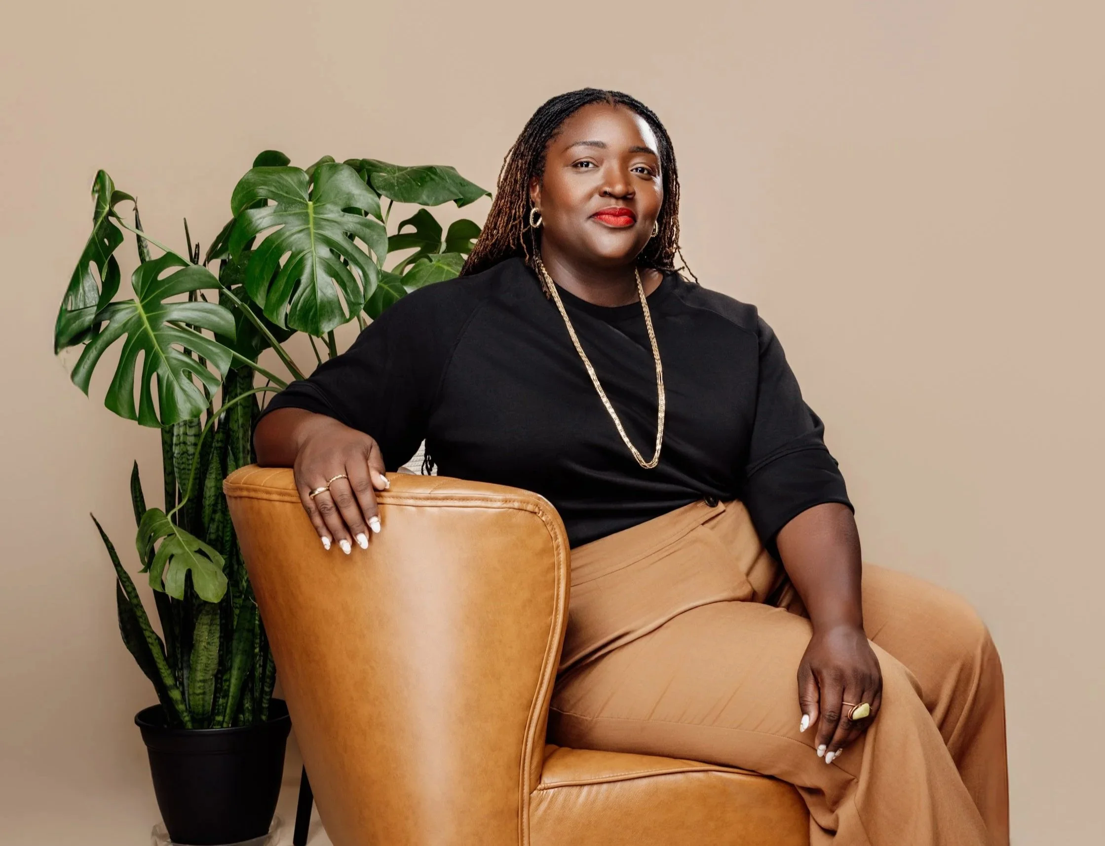 A beautiful and confident dark skined Black woman with locs poses sitting on a tan leather chair, wearing a black top, tan pants, gold jewelry, and red lipstick, next to large green houseplants against a beige background.