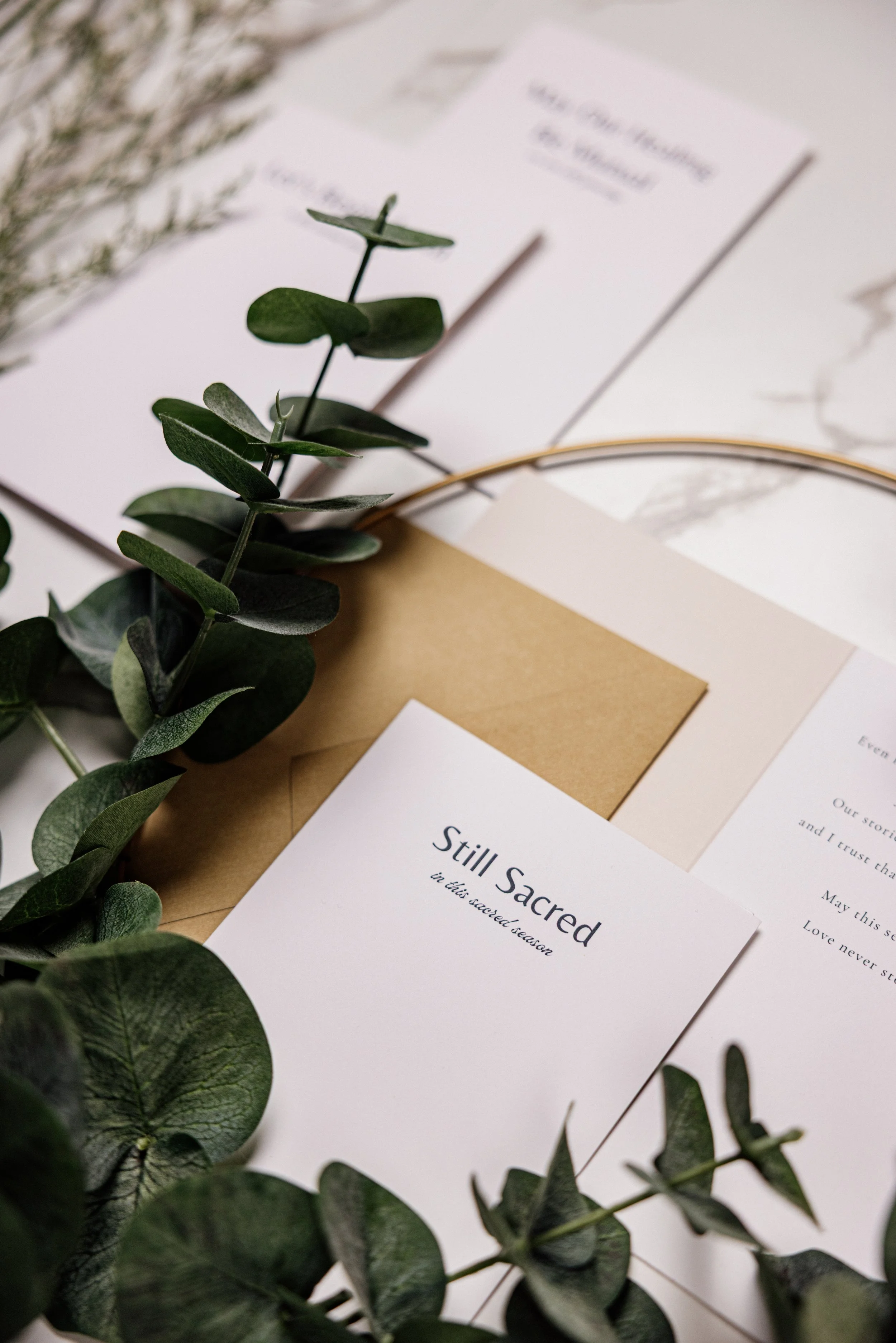 Close-up of a greeting cared with greenery. The visible card reads 'Still Sacred in this sacred season' with additional decorative paper and leaves around it.